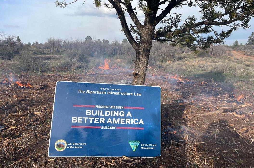 Image of a smaller ponderosa pine tree, small flames of fire, brush and wood chips with a blue sign against the tree with text saying, 'Project Funded by The Bipartisan Infrastructure Law Building A Better American Build.gov, US Department of the Interior and Bureau of Land Management'