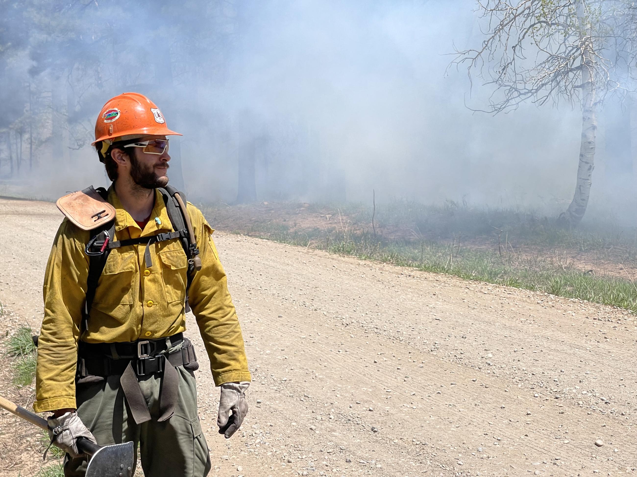 Firefighter with orange hard hat walks down the road with smoke in the background