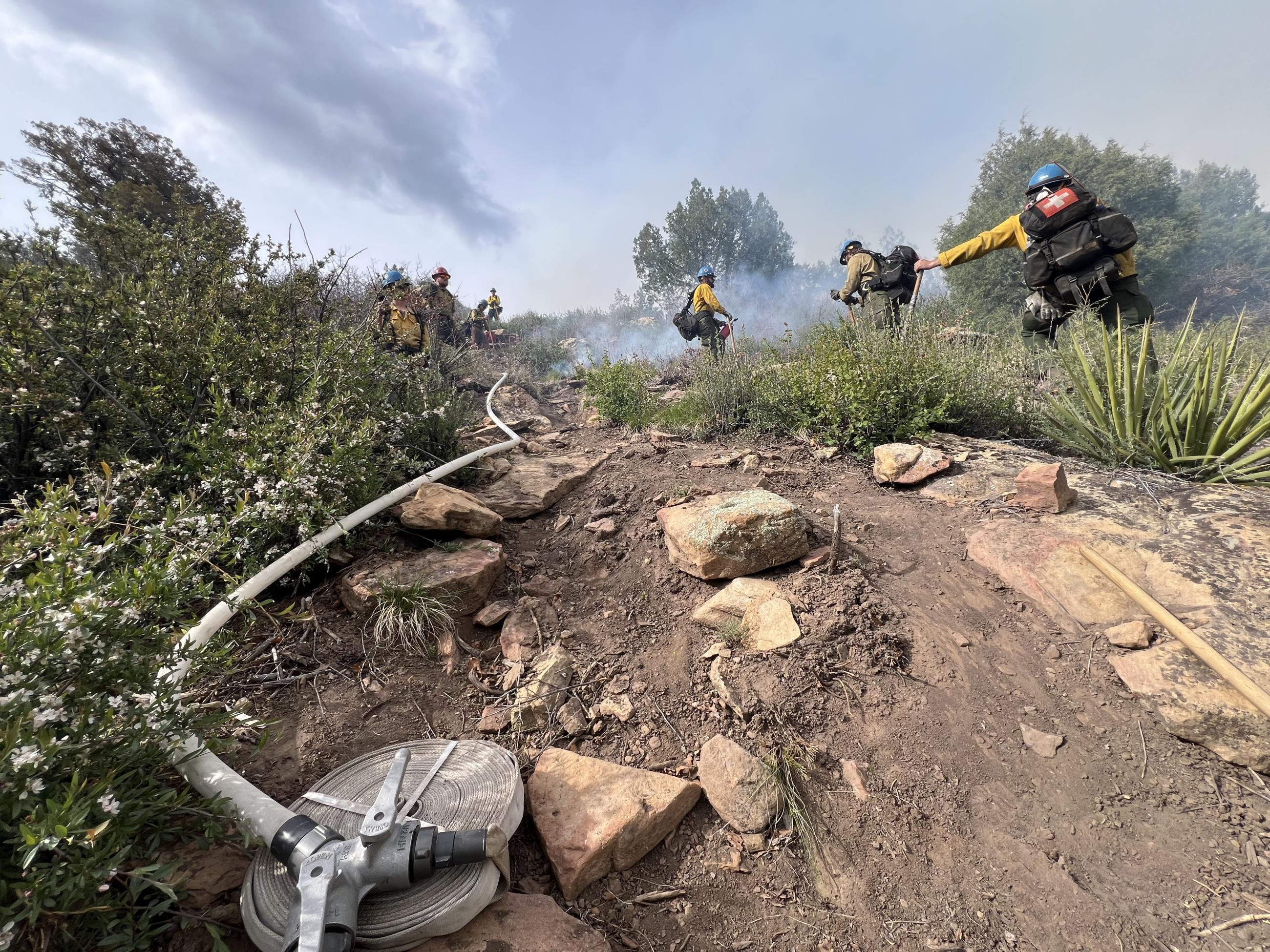 Firefighters are seen standing on a steep slope during a prescribed burn with a fire hose visible in the foreground.