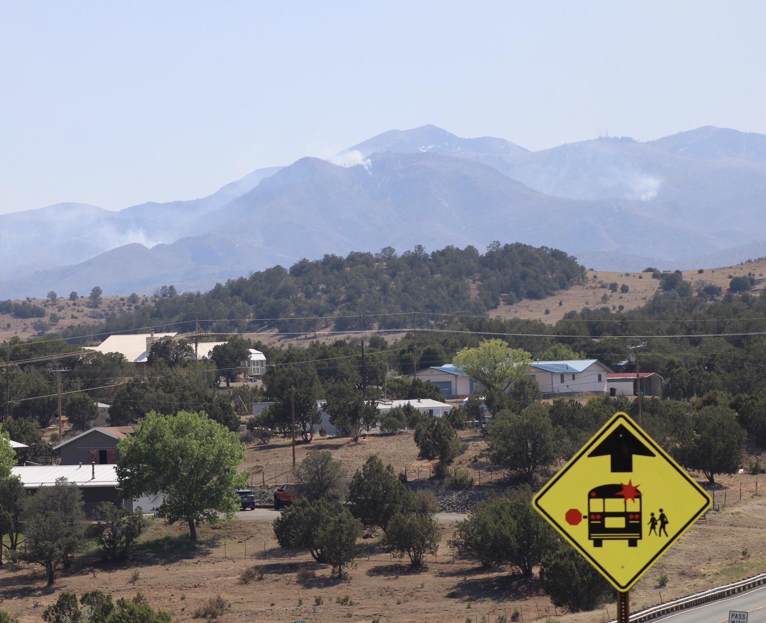 Houses and a school bus stop sign in the foreground are shadowed by smoke coming over a mountain range  