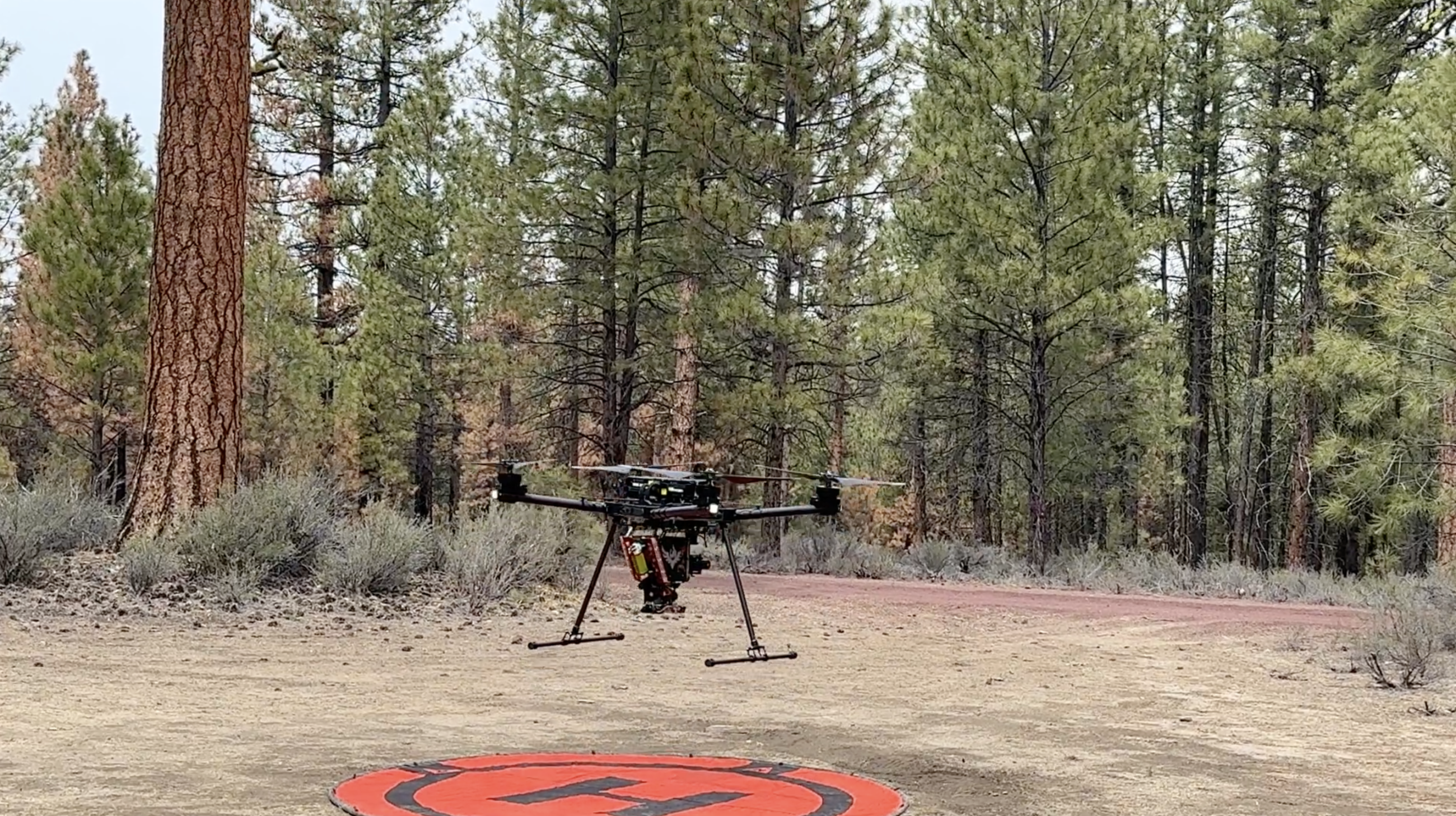A drone takes off from an orange H pad in the forest