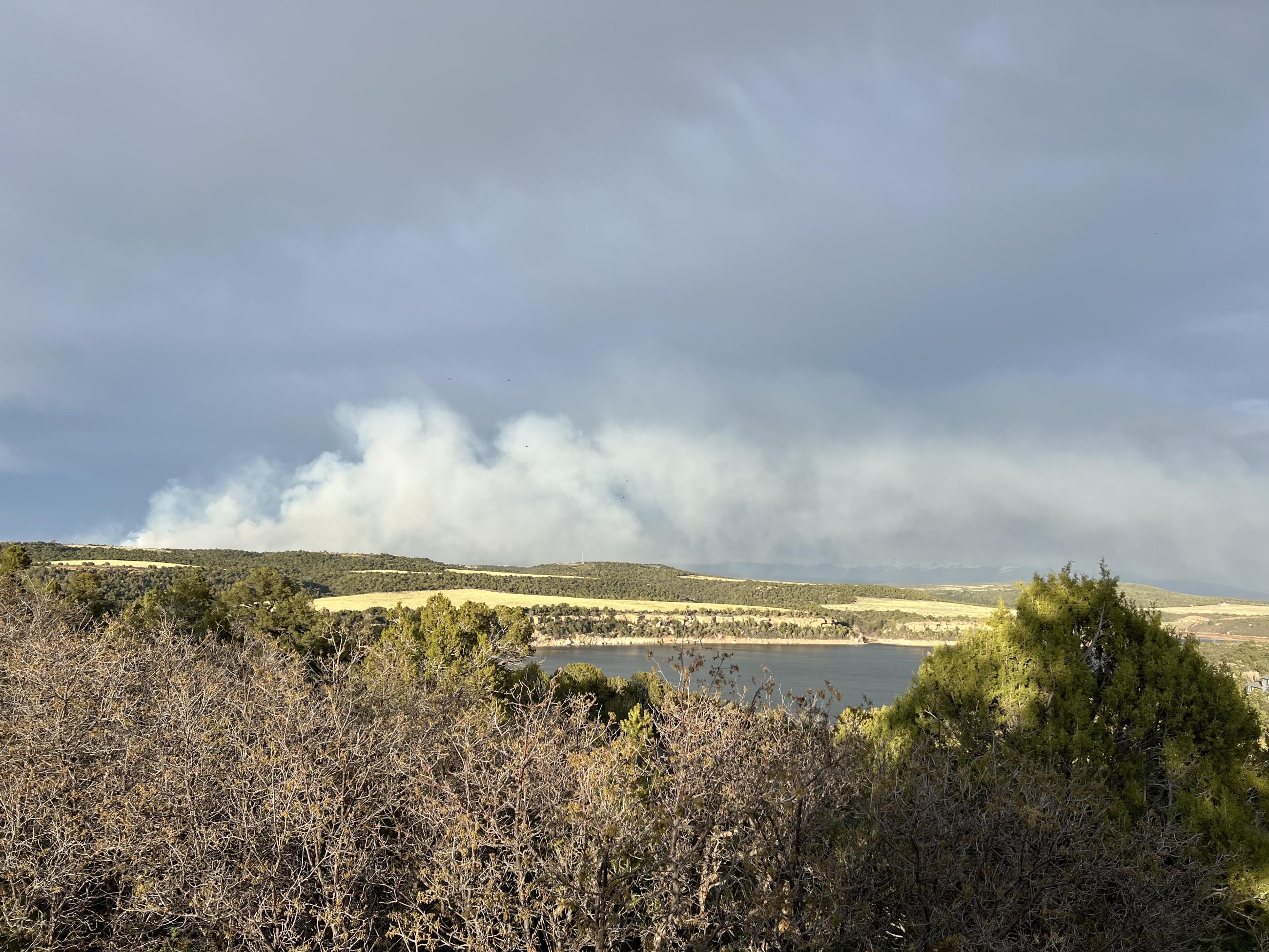 Smoke rises above the horizon and a portion of a reservoir is visible in the foreground.