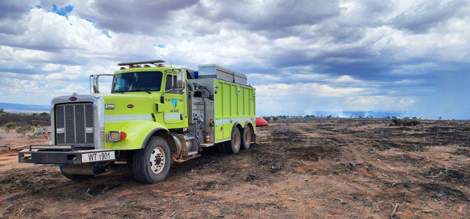 A lime green wildland fire fighter vehicle or water tender truck is parked on a desert landscape scorched black by a wildland fire. An orange "pumpkin" or mobile water dip tank sits in the background behind the truck.