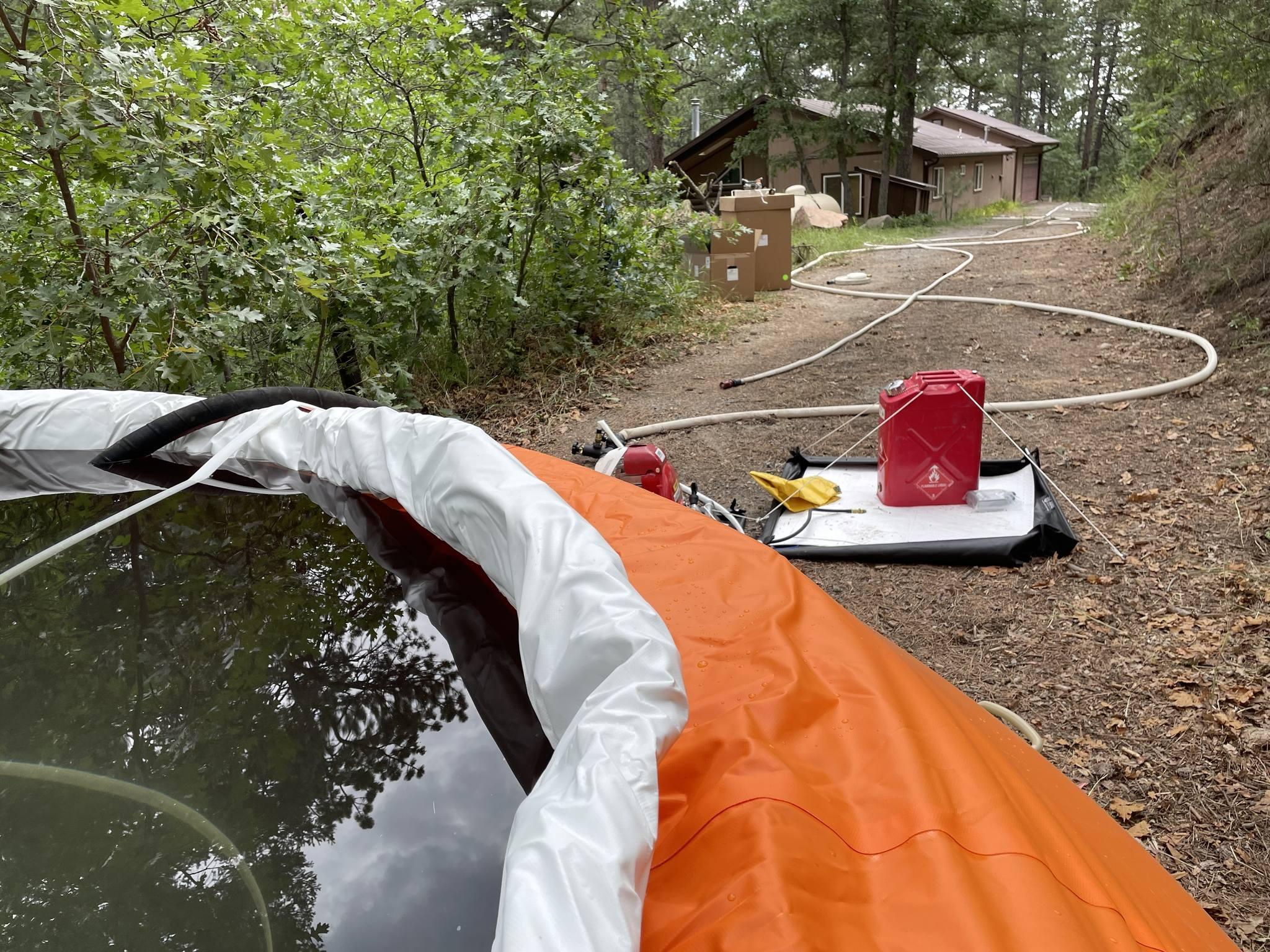 An orange water tank sits on the ground in the font of the photo. A red pump sits next to it, providing electricity to run water from the portable water tank to the hoses that are providing structure protection.