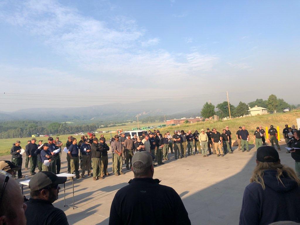 A row of firefighters stand in a parking lot for their morning briefing. The mountains behind them are hazy with smoke.