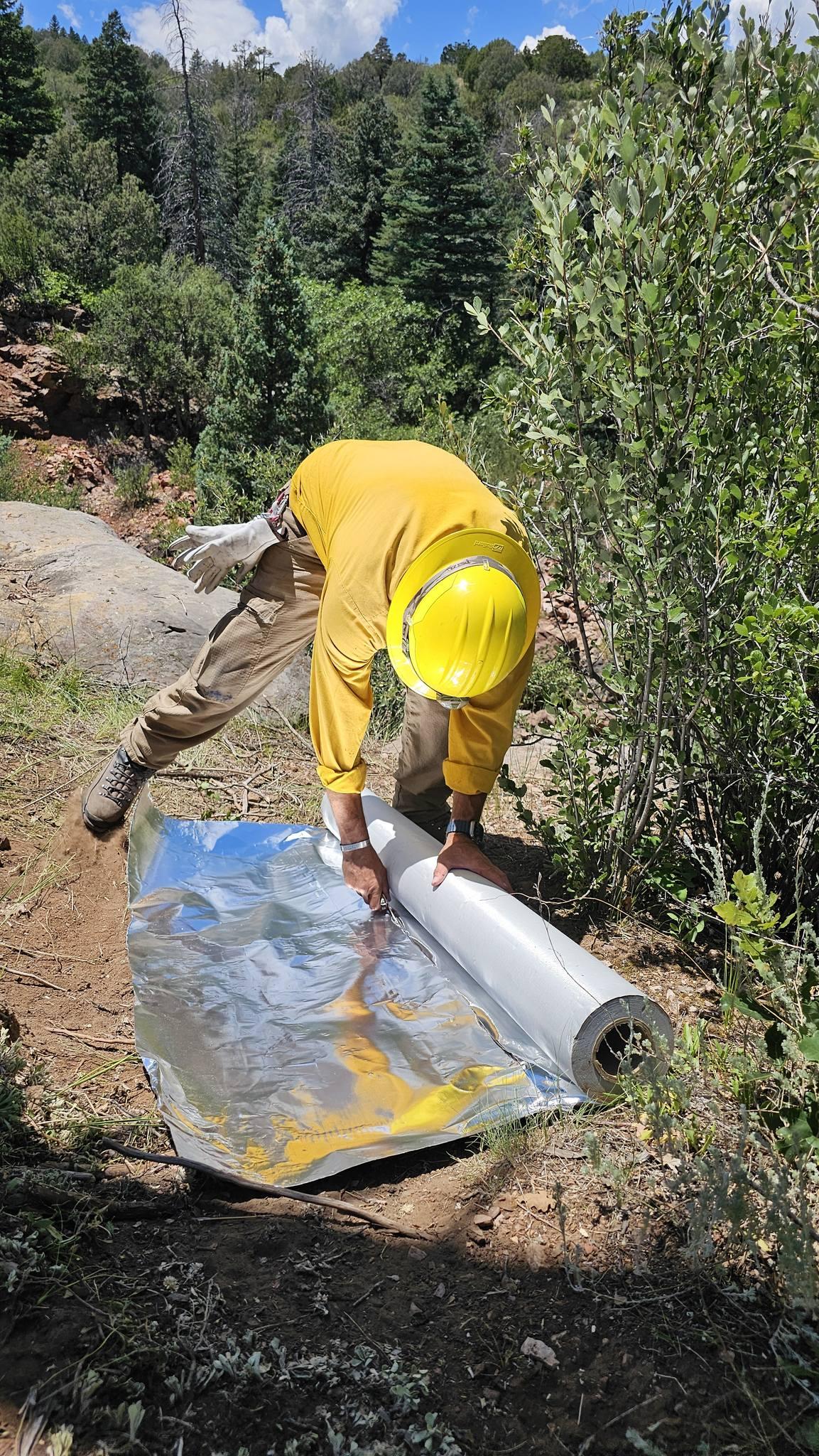 A firefighter in protective clothing kneels on the ground, unrolling silver protective wrap. 