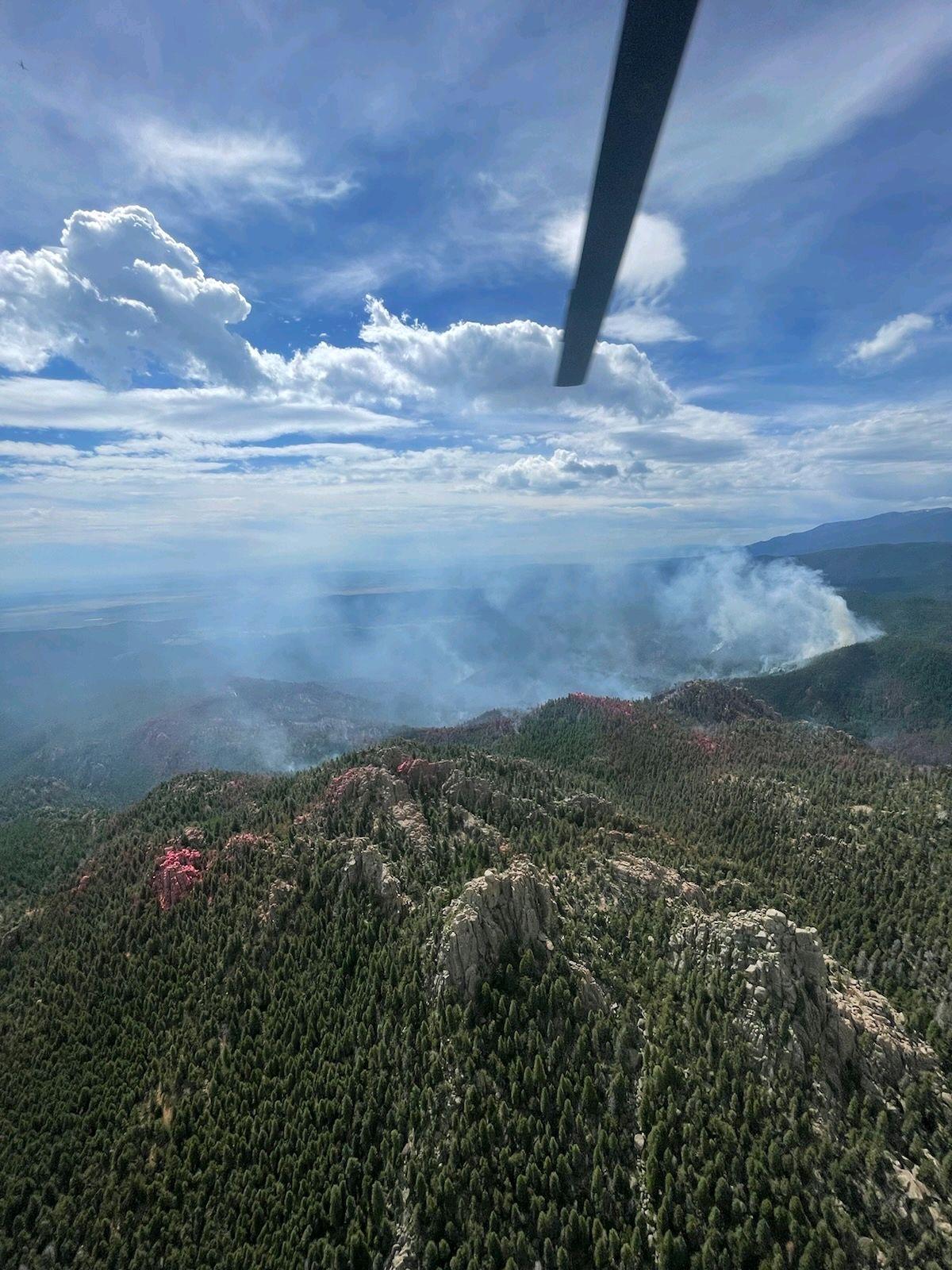 An aerial view of the fire. A helicopter blade is in the top of the photo, with a blue sky and fluffy white clouds above the ground. Smoke is drifting up in the middle of the photo. 