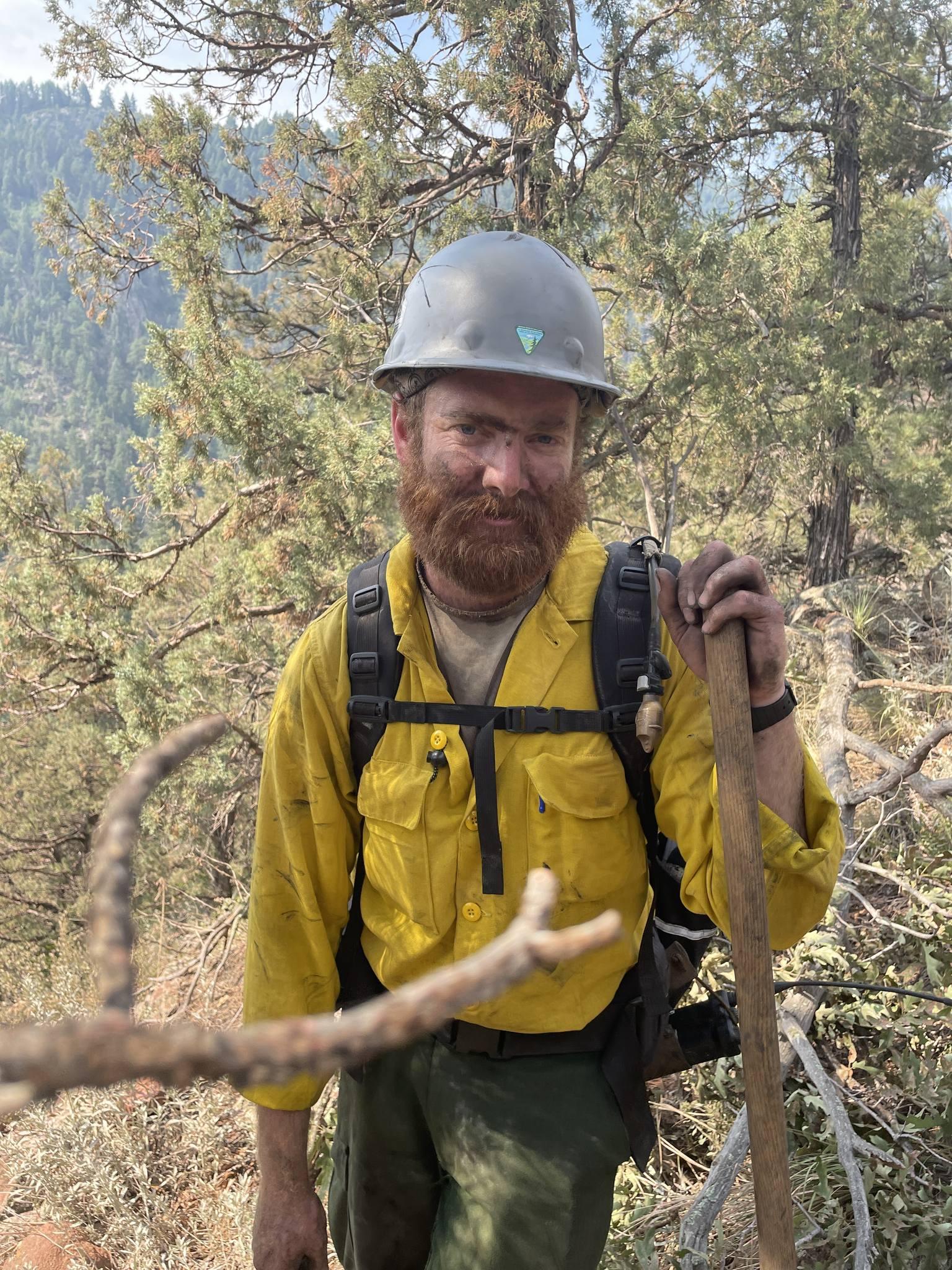 A firefighter in protective gear stands facing the camera. He has a silver hardhat on and the photo is from the waist up. A mountainous landscape with evergreens is behind him.