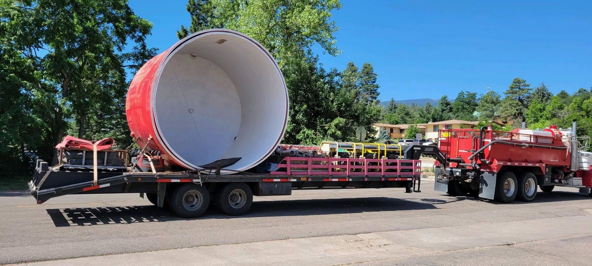 A heliwell is being delivered on the back of a trailer that is being pulled behind a truck. A heliwell is a portable water tank that helicopters can use to refill their water tanks.