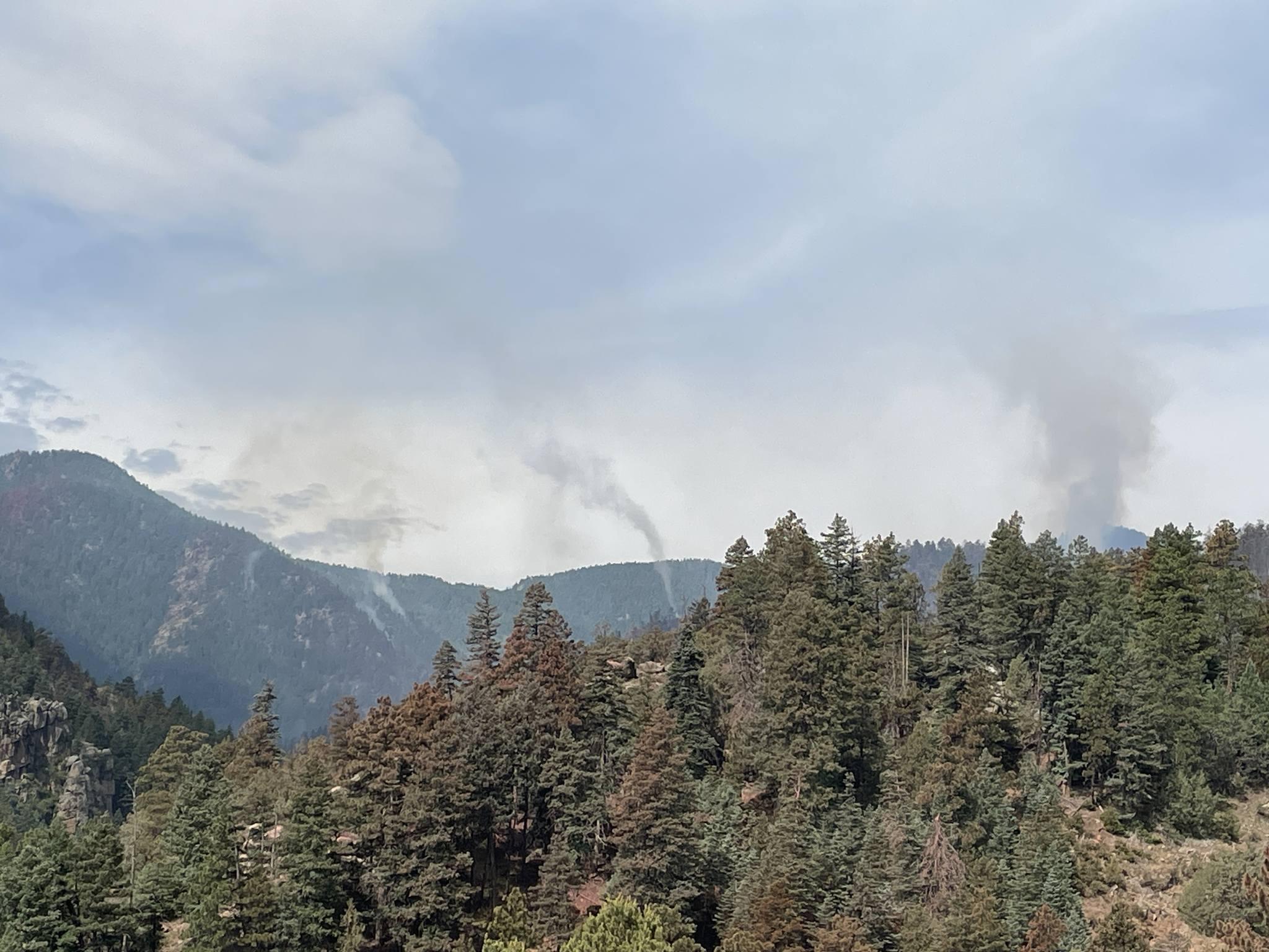 A wide view of smoke rising up from the fire. In the front, evergreen trees fill the photo, in the background, mountains with several small vertical columns of smoke rising towards a cloudy sky.