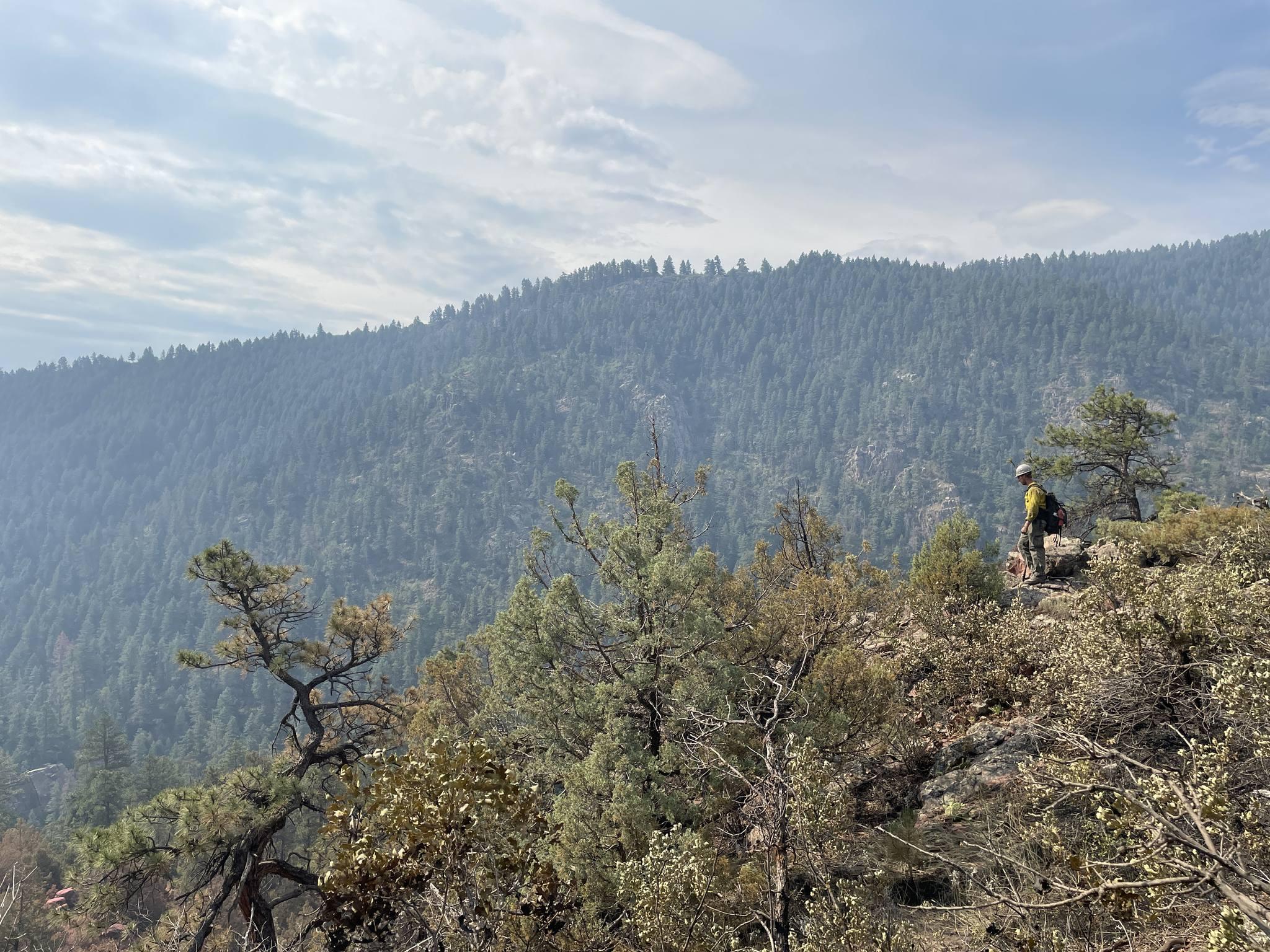 A firefighter in Nomex stands on the edge of a sparsely vegetated overlook of the Oak Ridge Fire. A mountain full of evergreens is in the background. Clouds almost fill the blue sky.