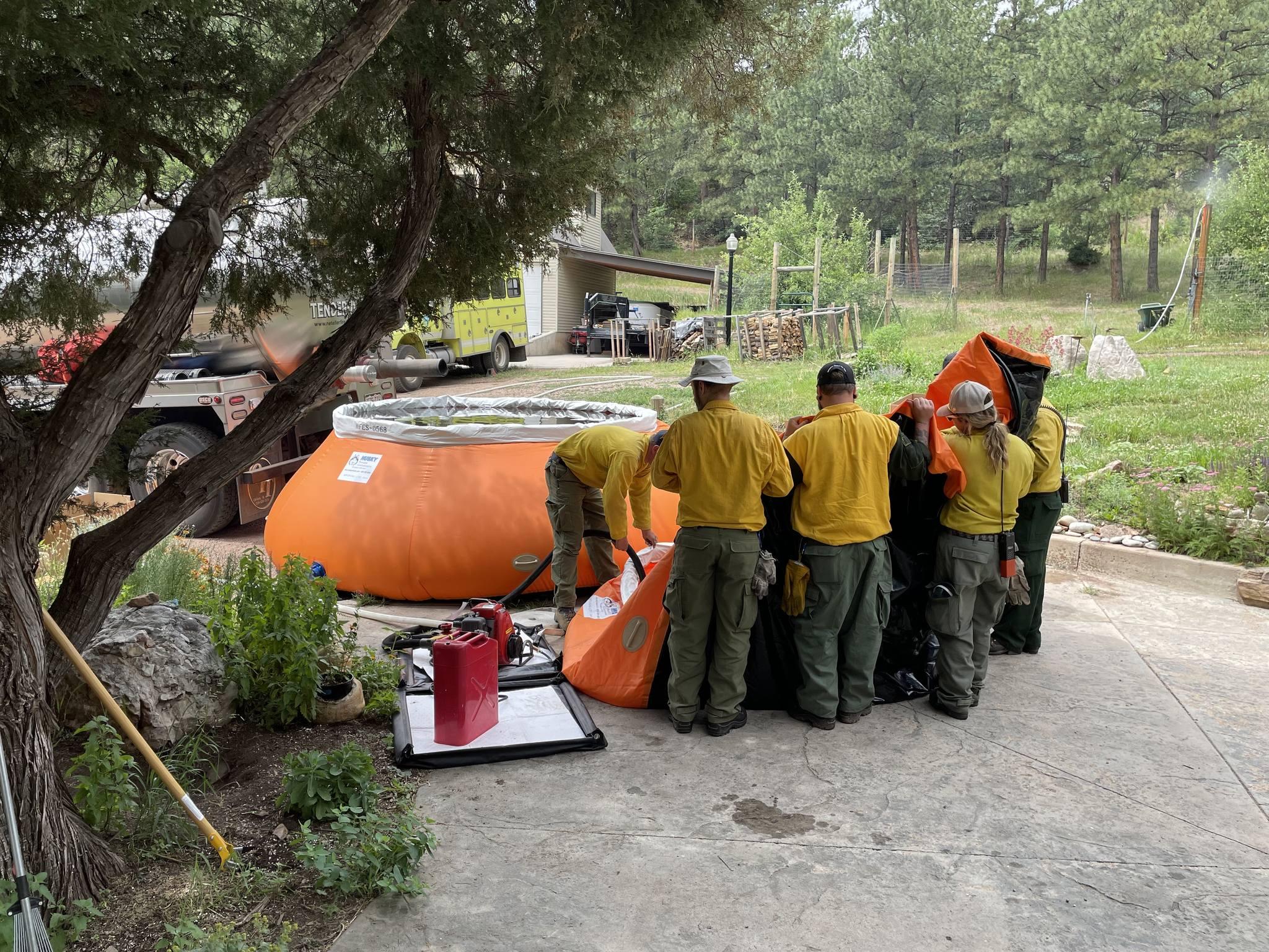 One orange water tank filled with water is towards the back of the photo. In the front of the photo, 5 firefighters are working to fill a new orange water tank.