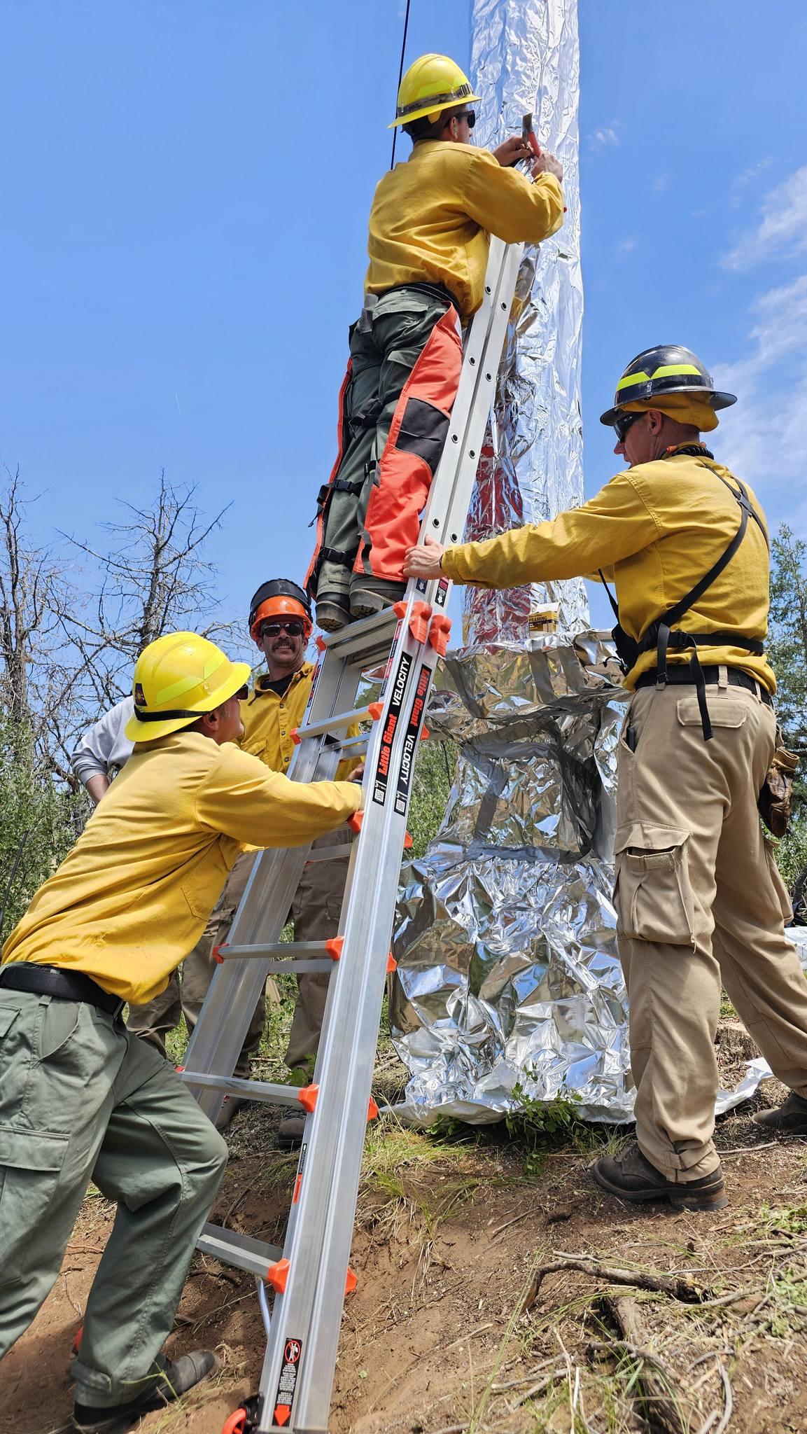 Three firefighters in Nomex are wrapping the derrik that was used to mine Beulah County Marble. One is standing on a ladder and two are on the ground, with one of them holding the ladder. 