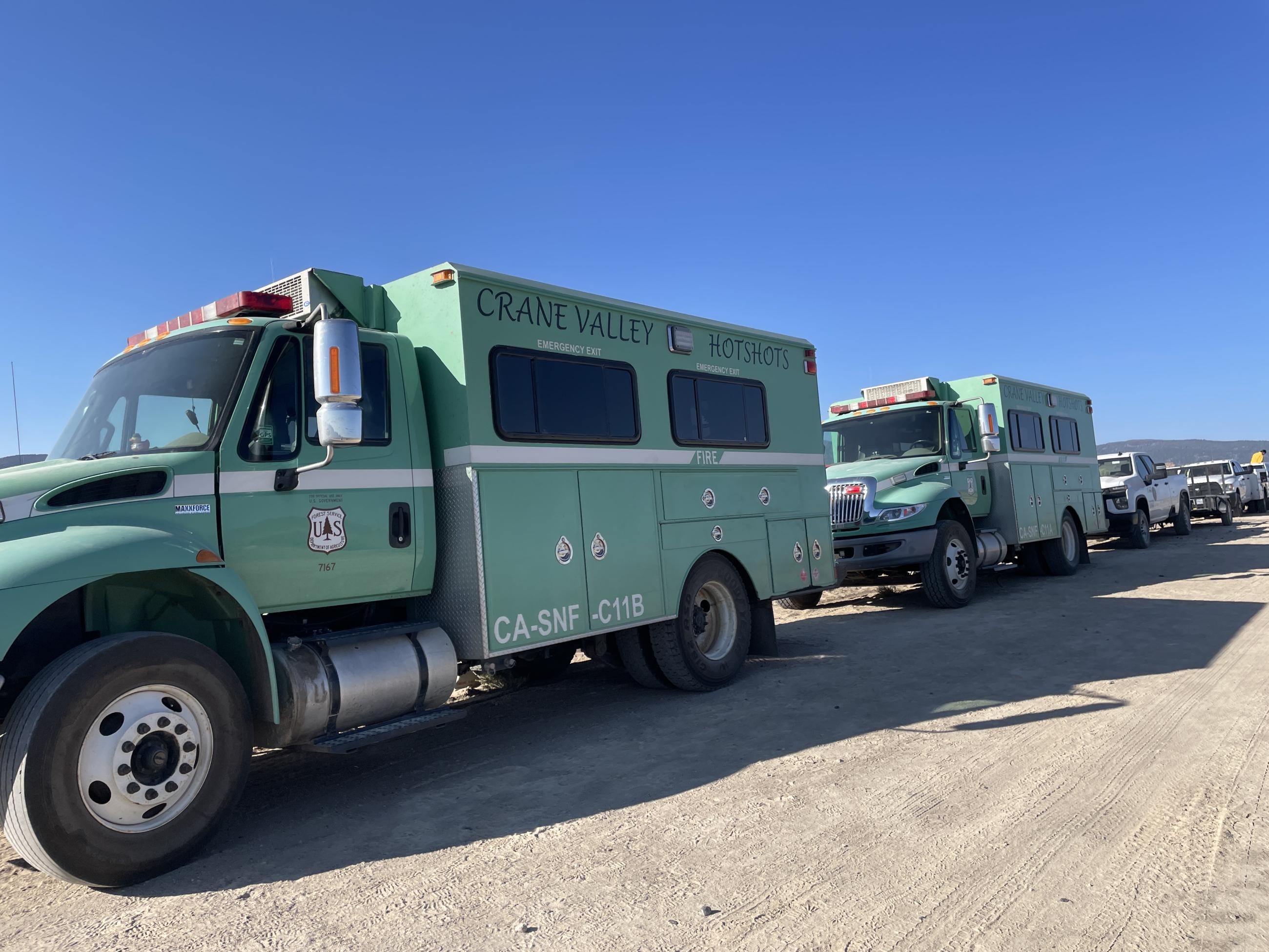 Two large, green, Crane Valley Hotshot crew trucks parked on dirt road with blue sky.