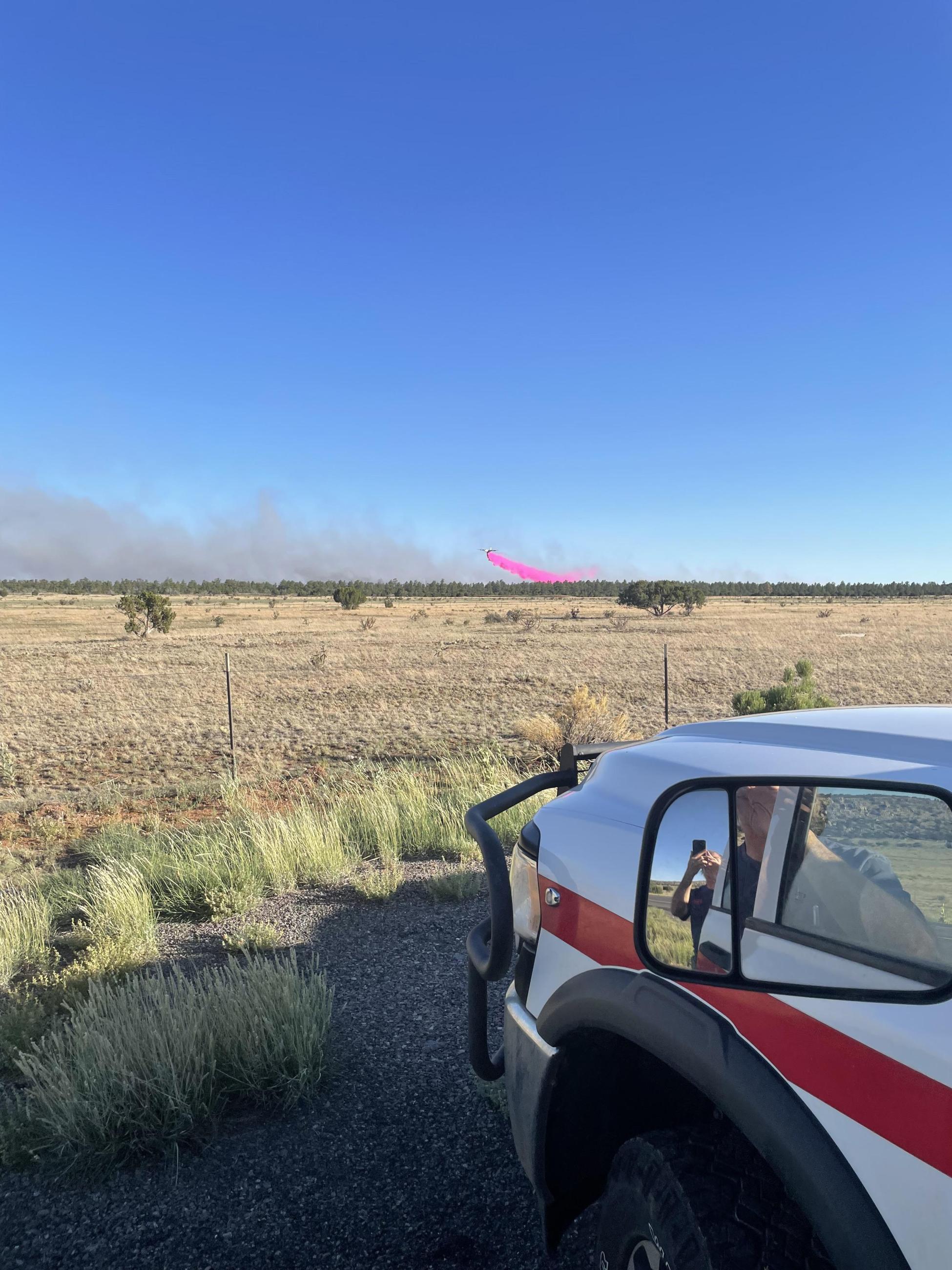 An air tanker lays down a line of retardant over the Encerrita Fire in El Malpais National Monument. The plane is barely visible but a long pink line of retardant is visible through the smoke. The photo is taken from a vehicle with grass along the roadside. 
