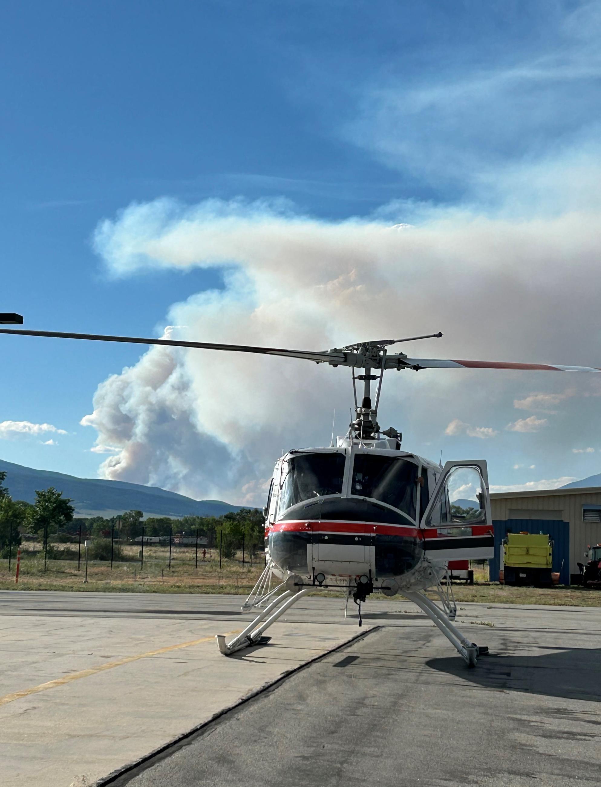 Photo of a helicopter assigned to the Interlaken Fire seating on a paved surface with smoke from the Interlaken Fire in the background. 
