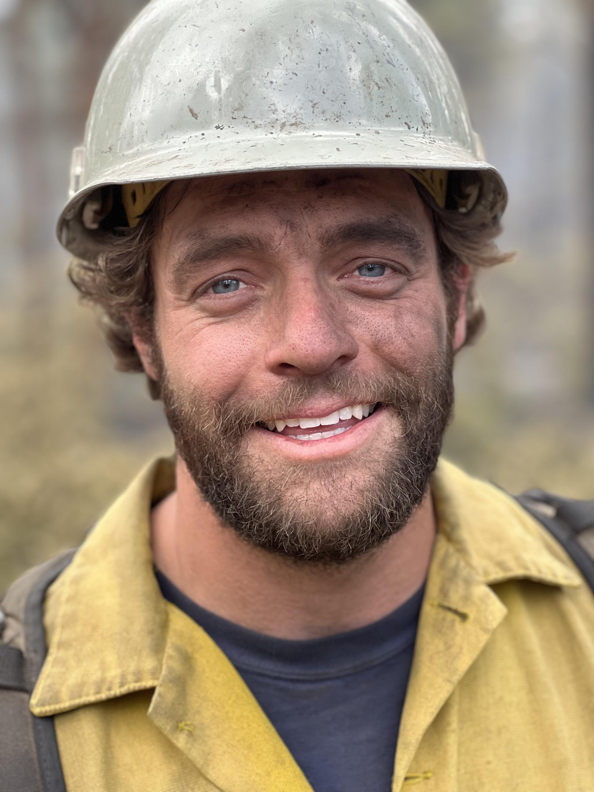 A bearded firefighter in a grey fire helmet smiles at the camera.