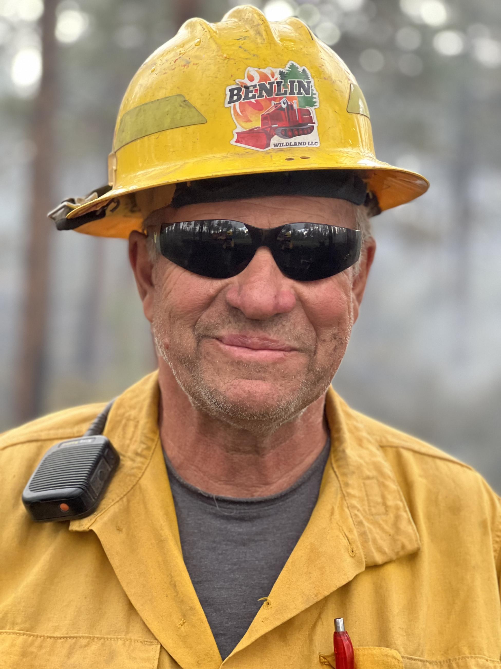 A heavy equipment operator in sunglasses, yellow fire shirt and yellow fire helmet.