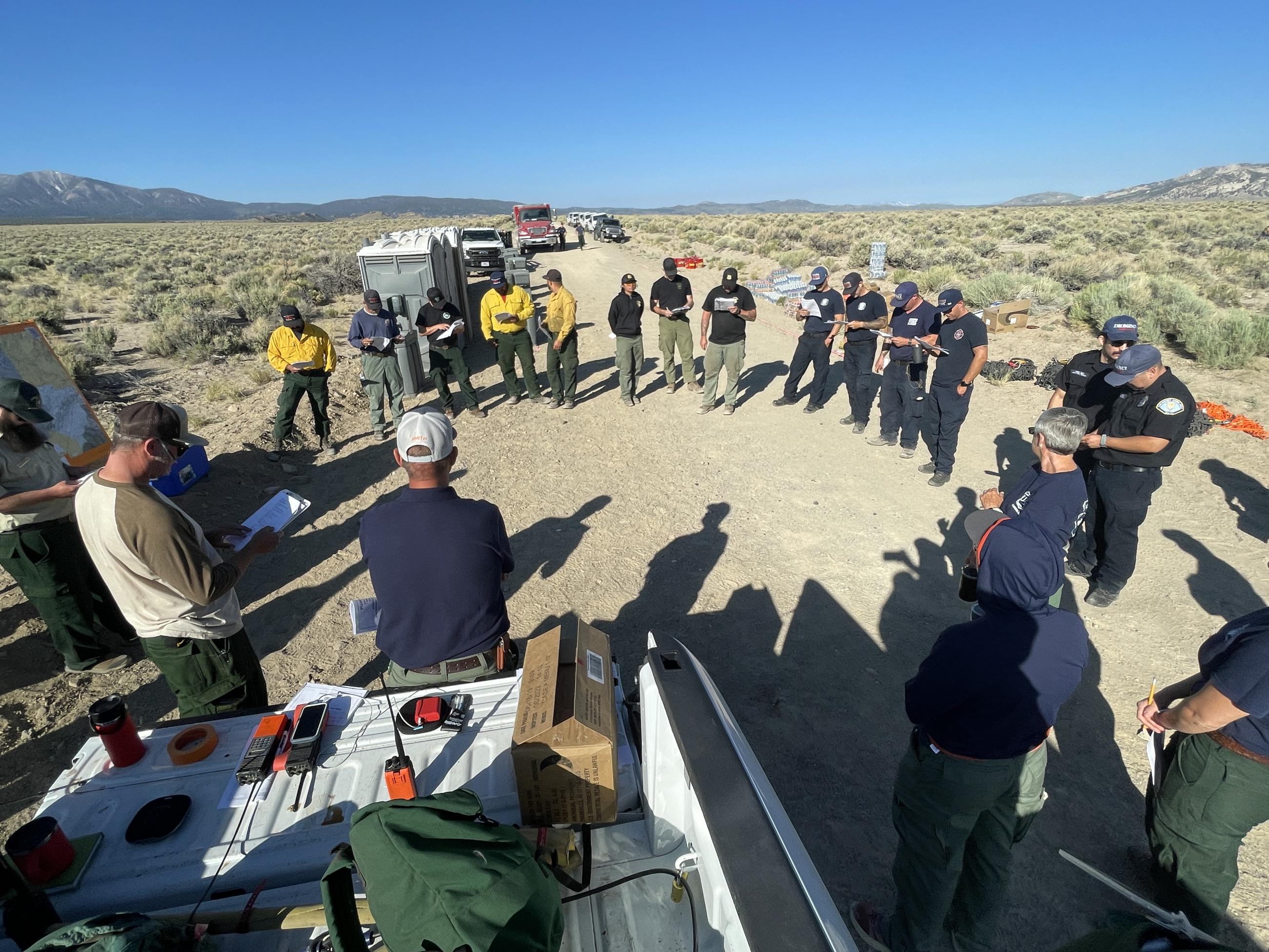 Fire personnel circled up on dirt road bordered by sagebrush under blue sky.