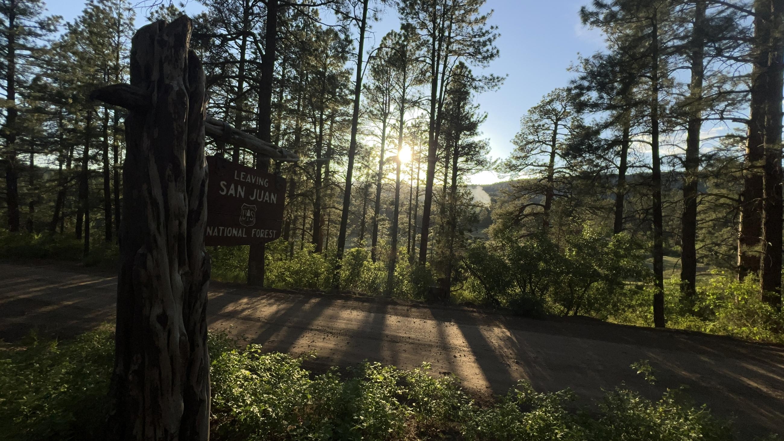 A San Juan National Forest entry sign is at left. The sun is setting behind pine trees and a smoke plume of smoke is at right.