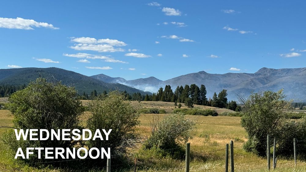 Blue skies, mountain with some white smoke in the distance, farm pasture with brush in the foreground.