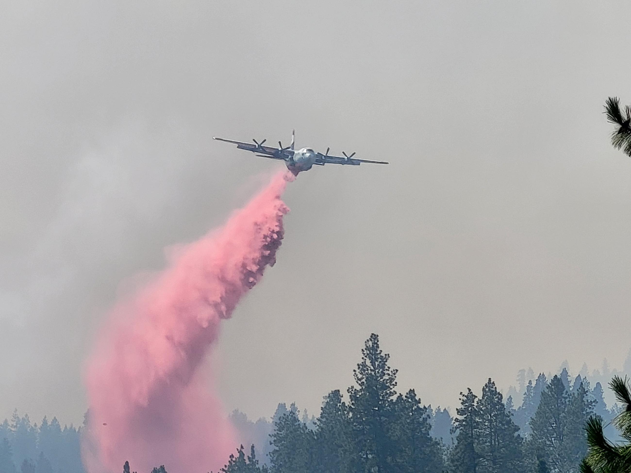 A plane flies towards the viewer in a smoky sky. Red retardant is dropping from the back of the plane