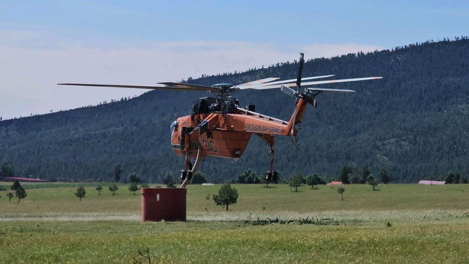 A helicopter is low to the ground, a hose comes out of the bottom of it. The hose is sucking water up from a portable well that is on the ground. 