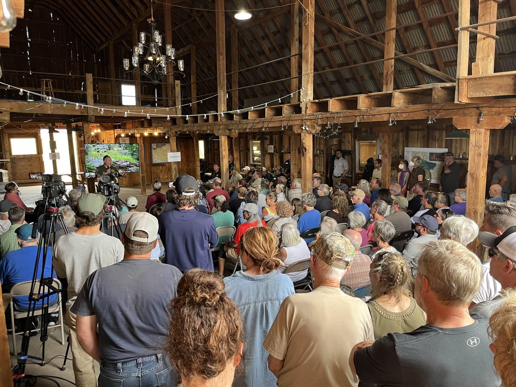 This is a photo of a public meeting that was held in the Goodplesant Barn. A rustic barn is packed full of community members, standing and sitting in chairs. At the front, a large screen shows presentation materials from the speakers.