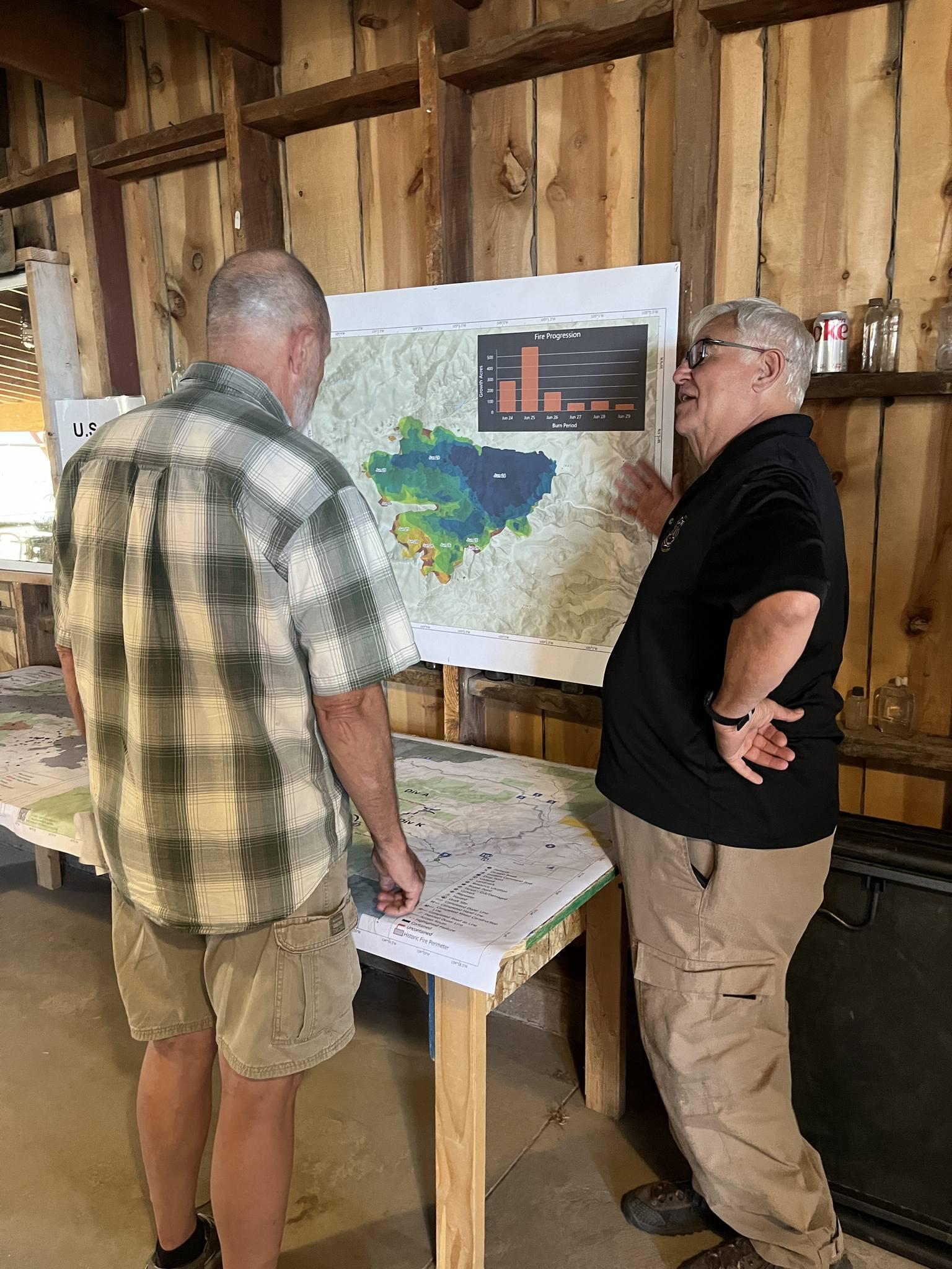 Photo is of the discussion after the community meeting. A US Forest Service representative stands next to a table. On the table and on the wall are maps of the Oak Ridge Fire. He is talking to a member of the public about the fire.