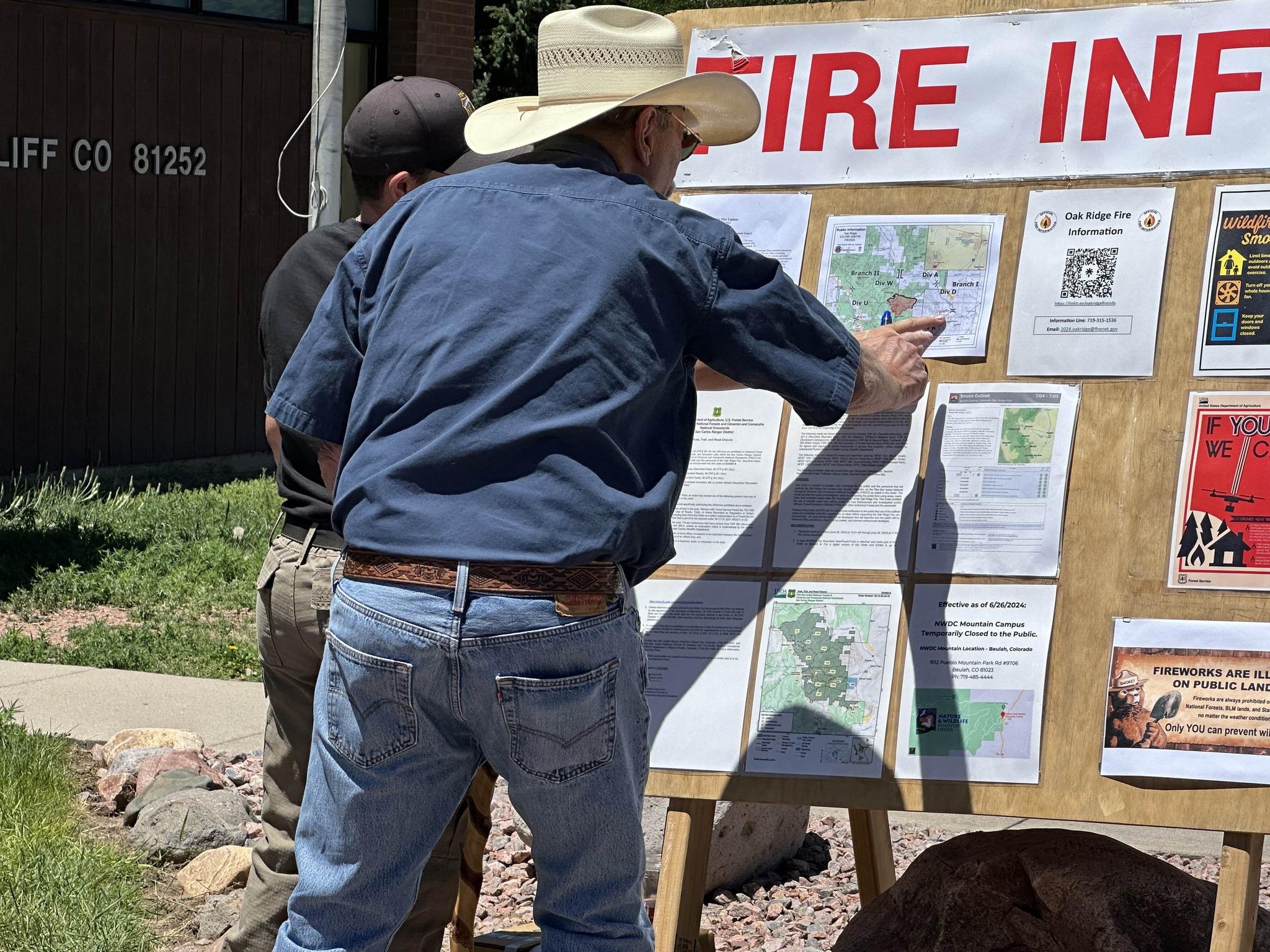 A man in a blue shirt and cowboy hat points to a fire information board that is being staffed by an Oak Ridge public information officer. 