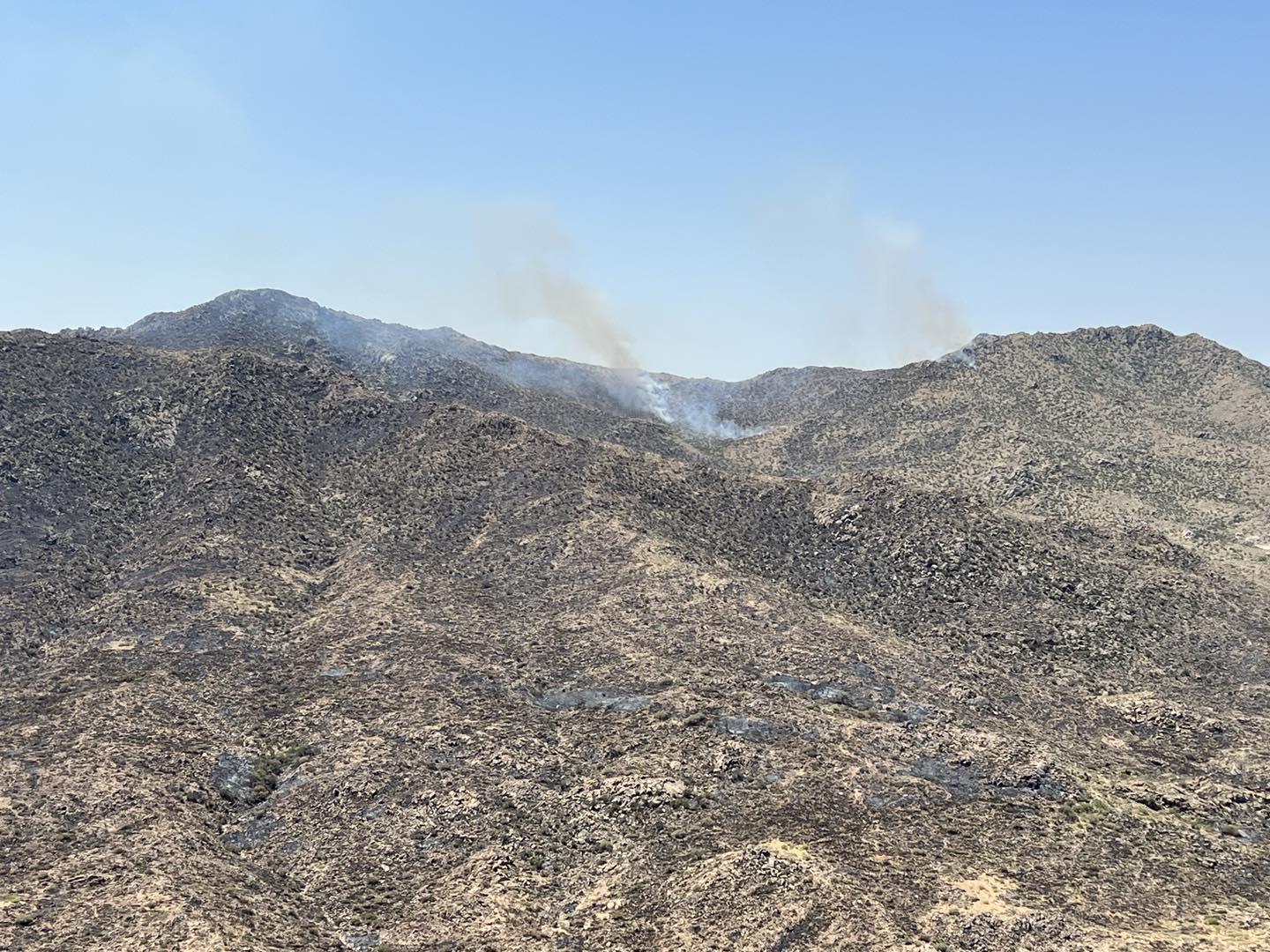 Mountain side with partial burned area. 2 light smokes columns rising against blue sky