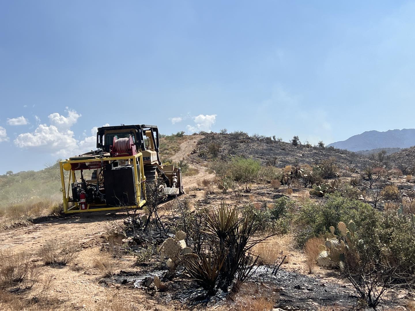 Dozer drives up dirt two track road with burned ground on one side, green vegetation on other