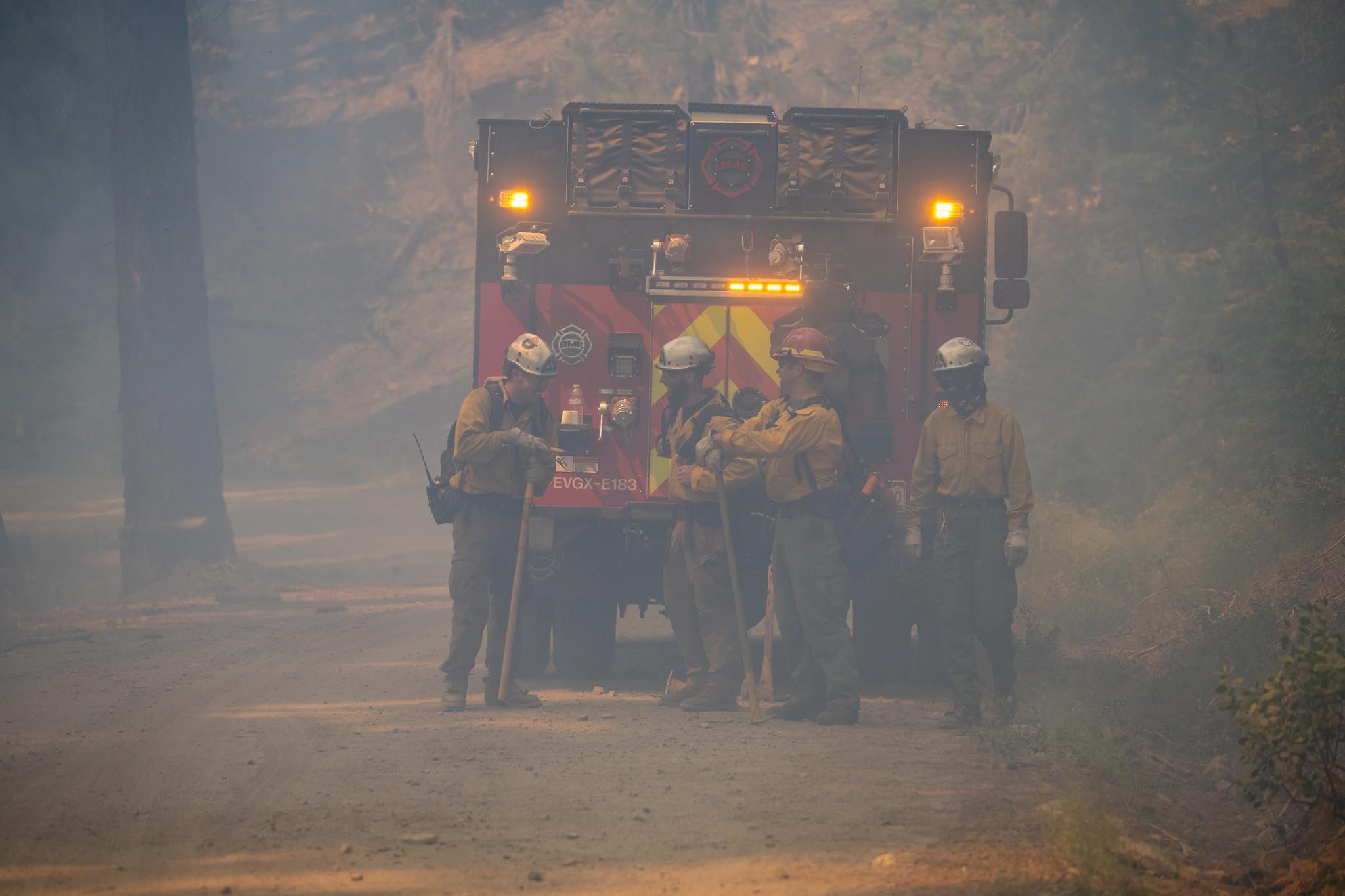 Four firefighters stand near an engine in smokey conditions on the Sheering Fire on July 6, 2024.