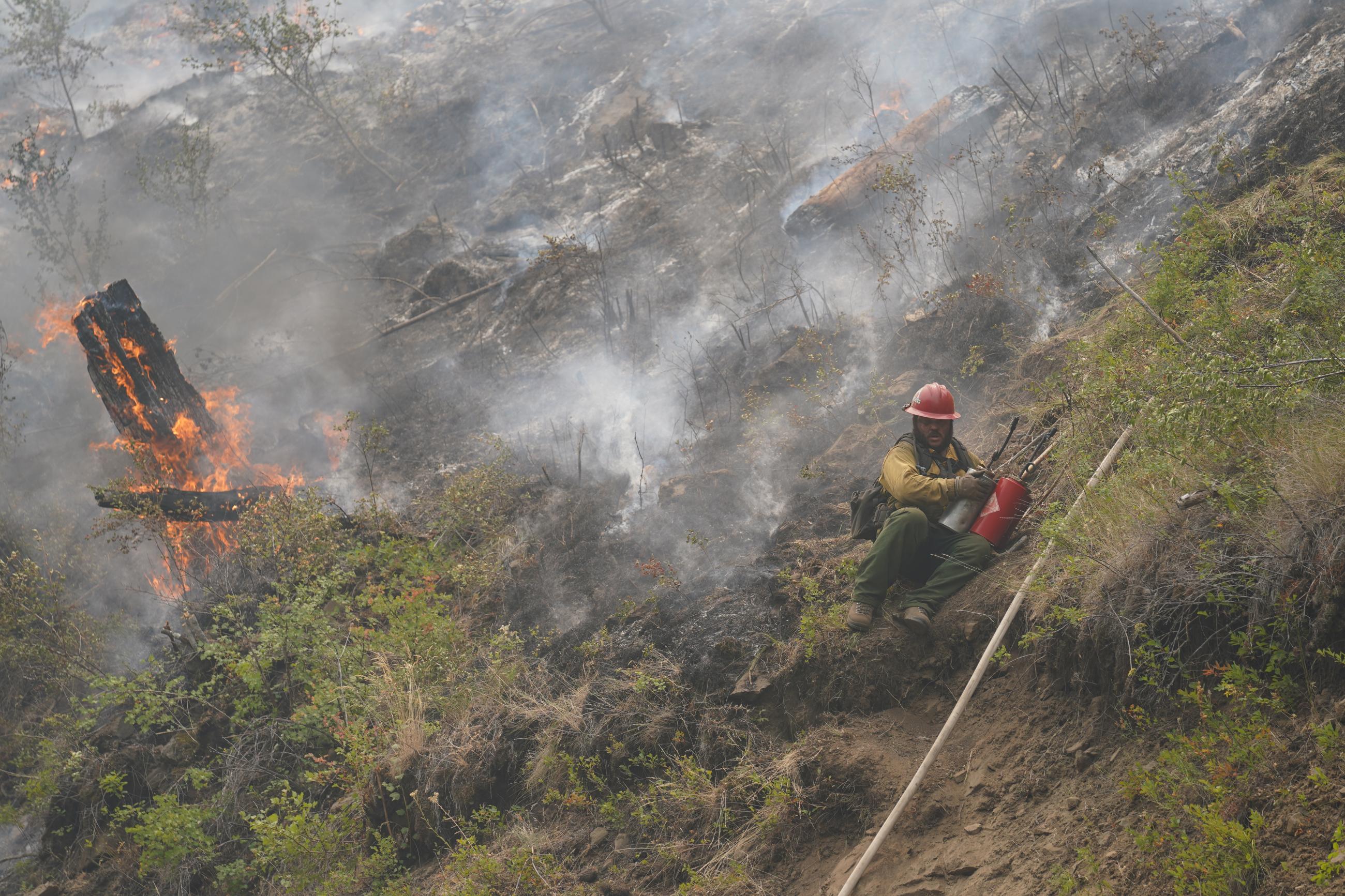 A wildand firefighter sliding down a steep hillside with oil cans in his hands and a burning stump in the background.