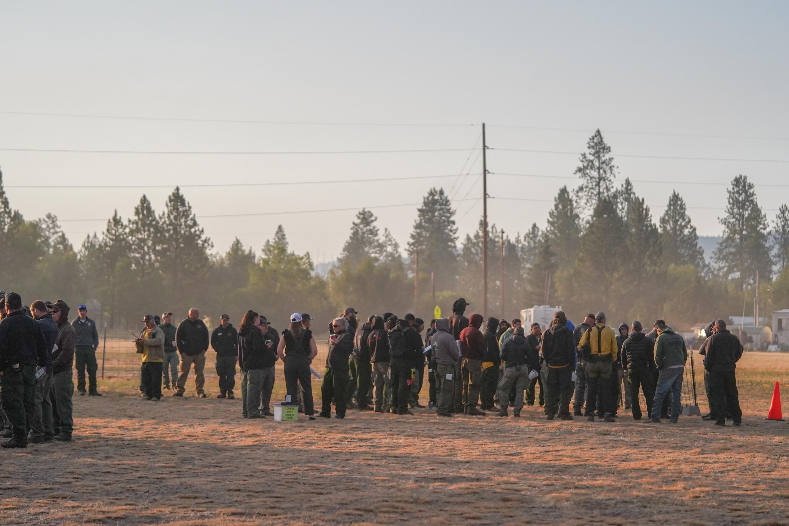 Image of dozens of wildland firefighters standing in a circle in a dusty field.