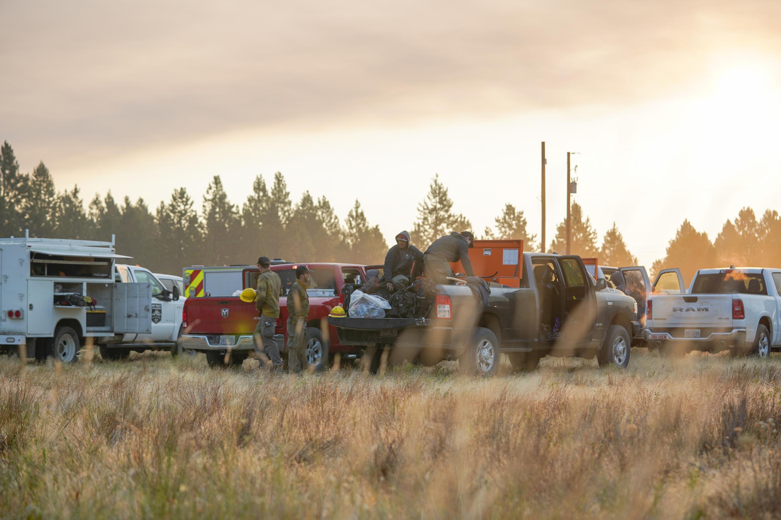 Sunrise behind firemen getting their gear ready in their trucks in a field.