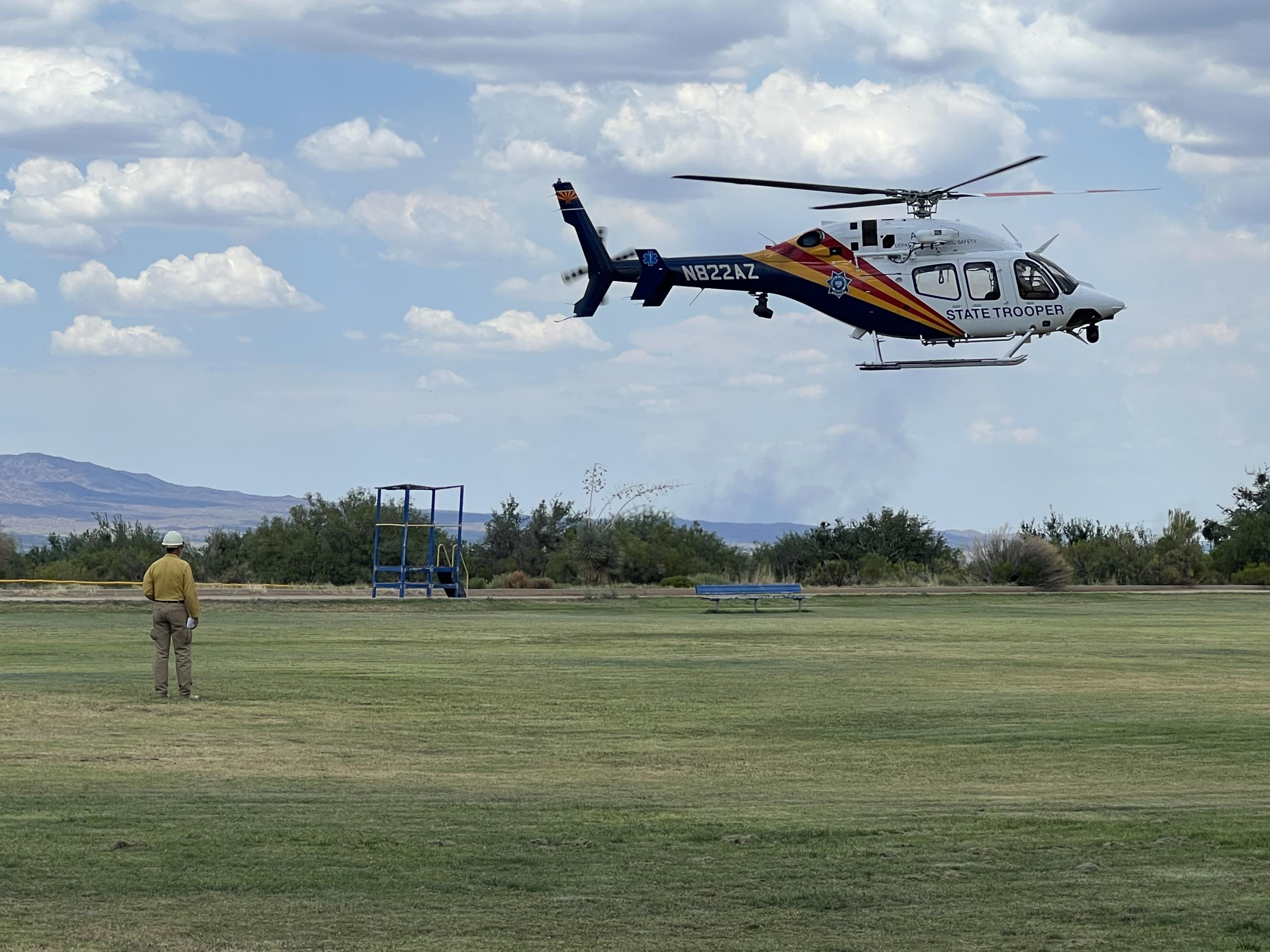 Helicopter landing on grass football field. Smoke column rises in the background