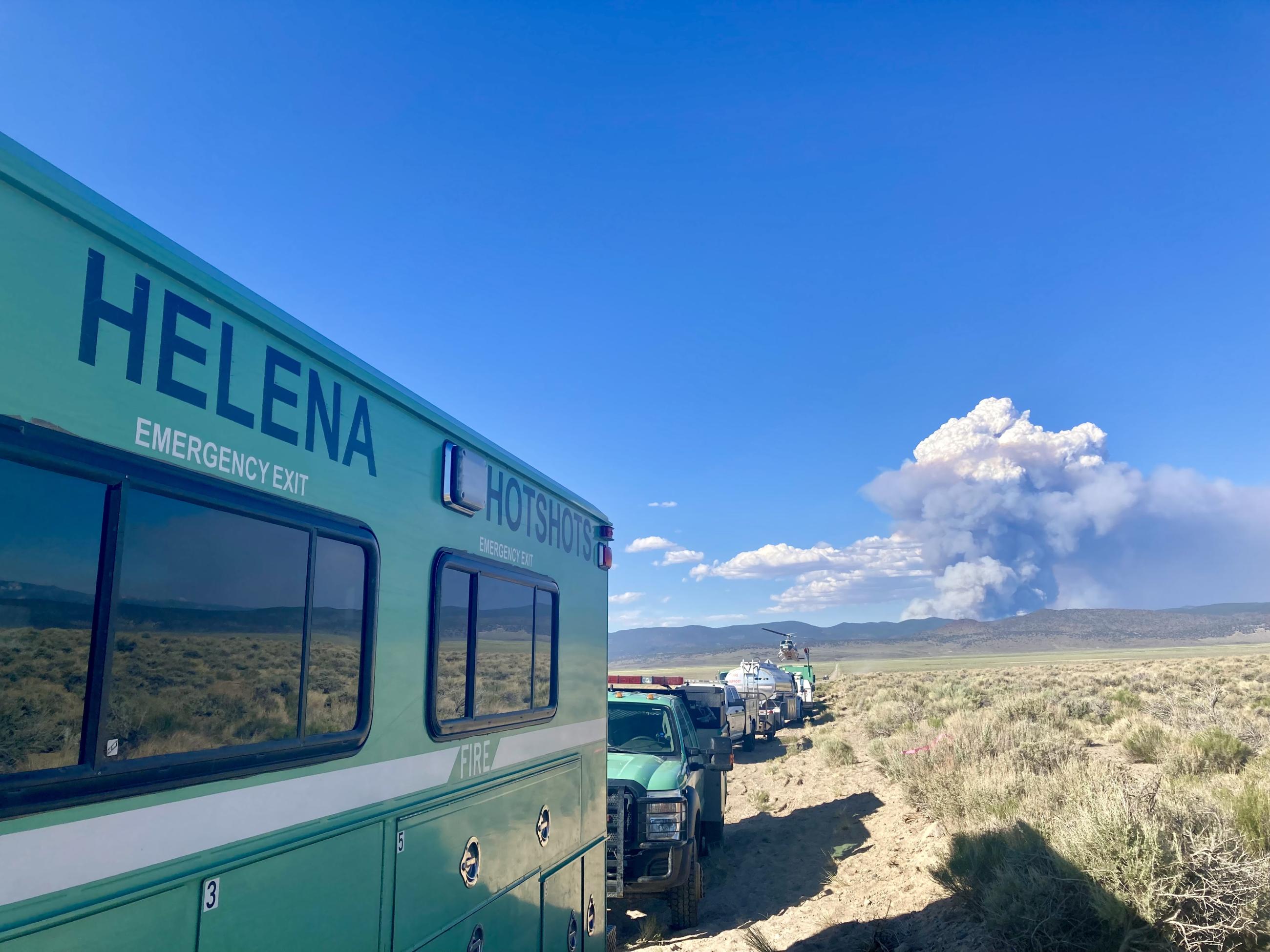 Large, green, trucks for the Helena Hotshot Crew on the road with smoke plume in the distance across the desert shrubs.