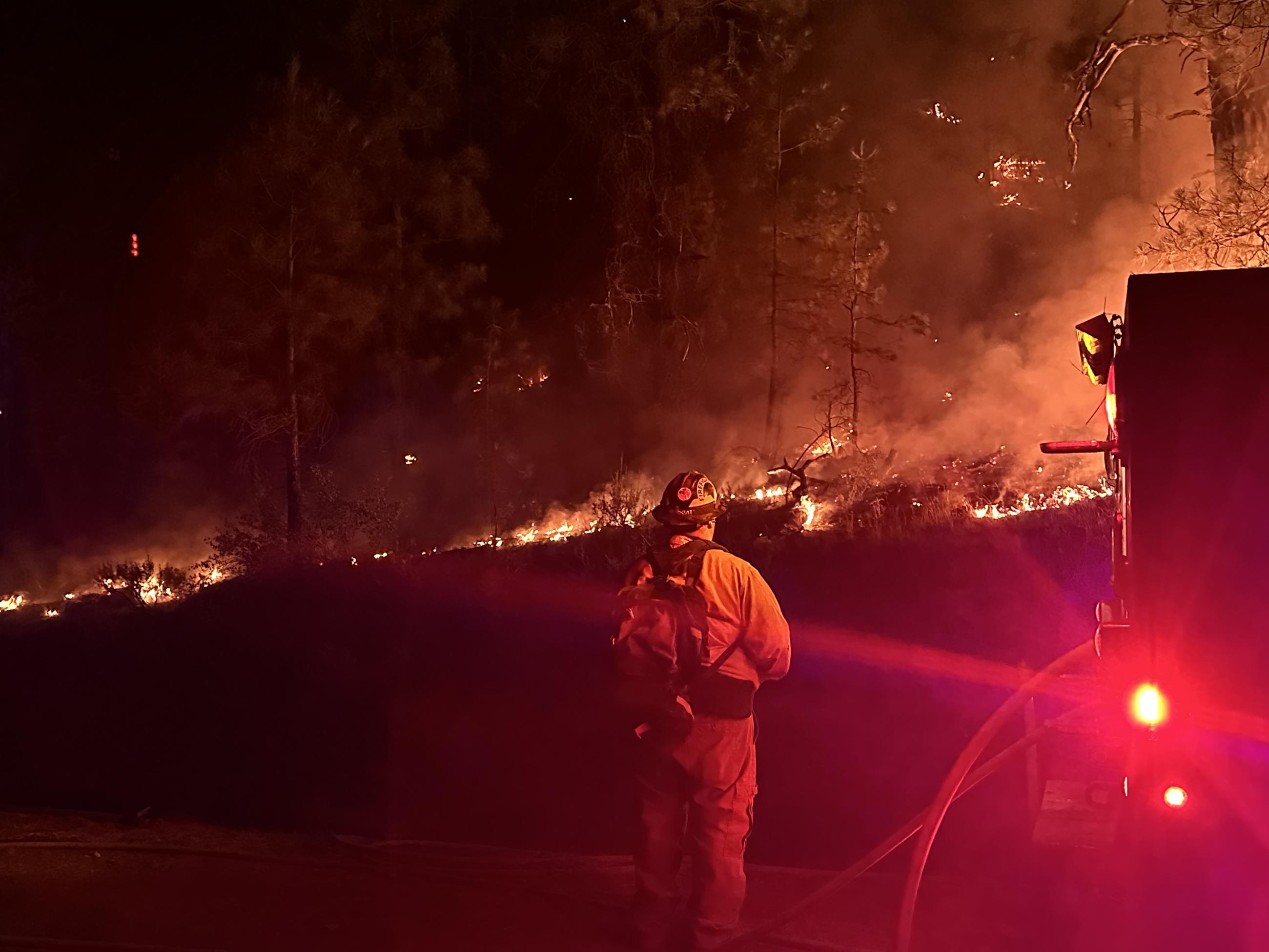 Night time, firefighter standing next to engine looking at flames and smoke