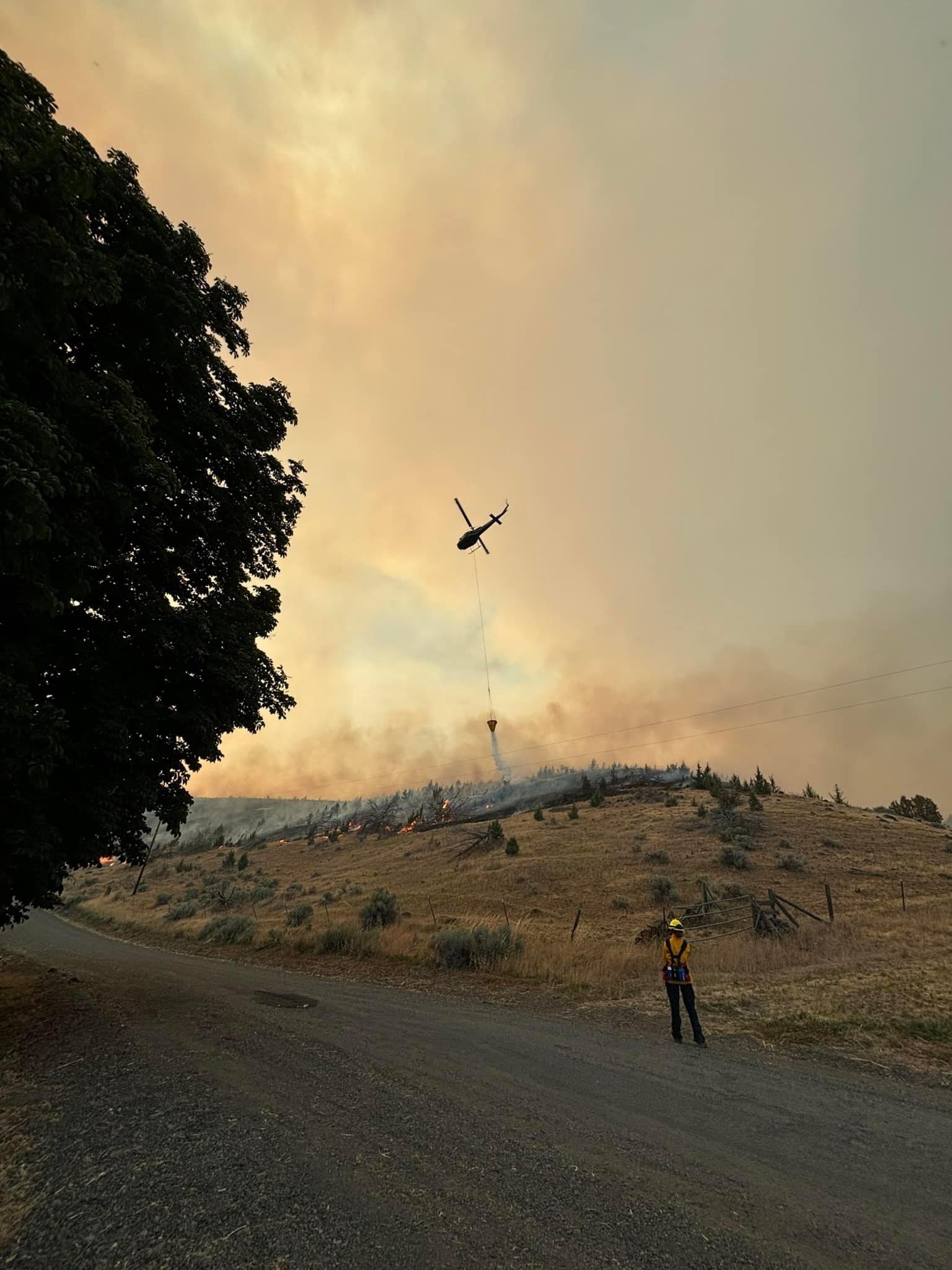 A helicopter drops water on the Lone Rock Fire with a firefighter standing on the roadside