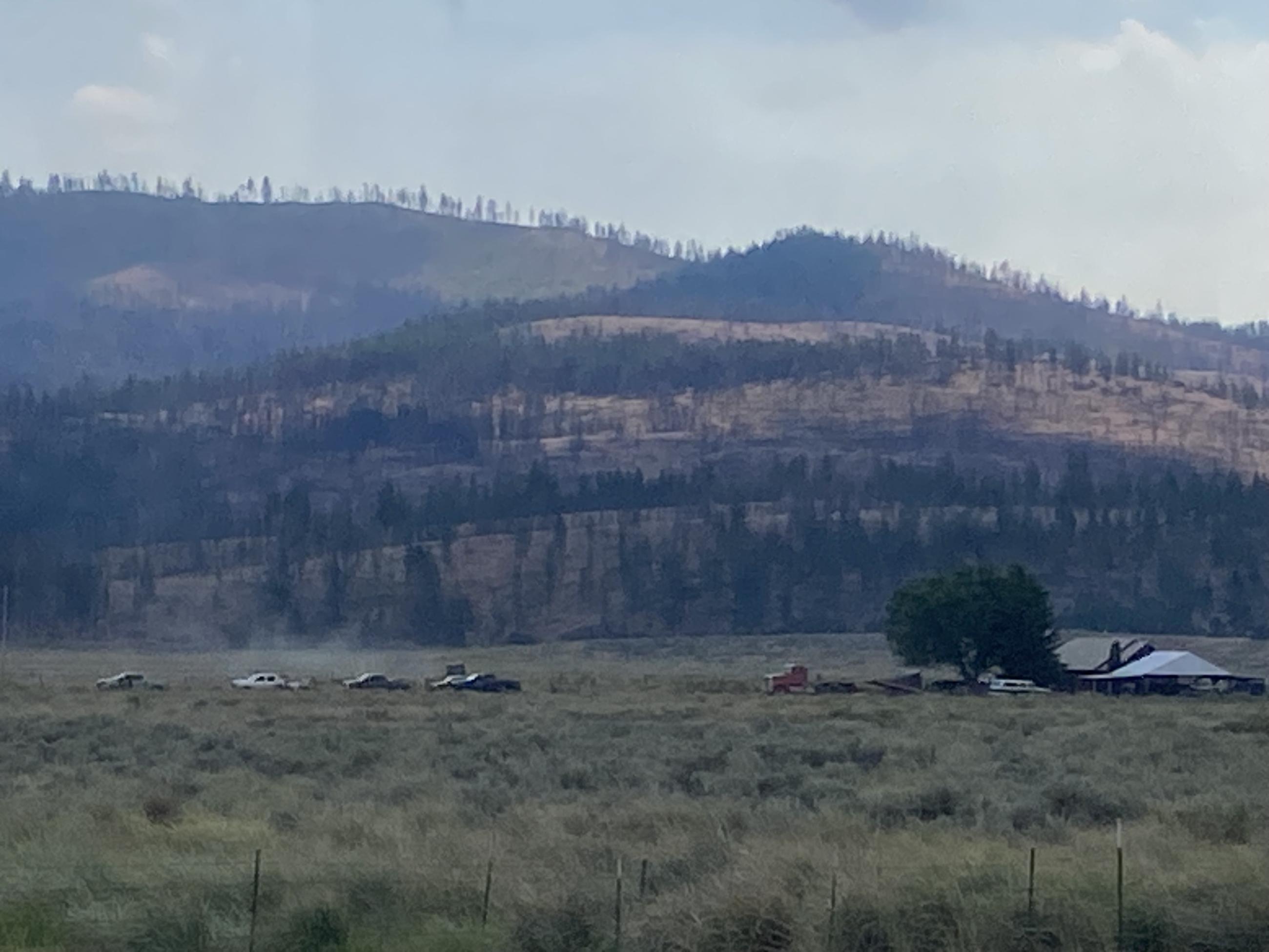 A field of sagebrush and grass with a home. Fire engines are driving to the home to start preparing the home for a possible wildfire.