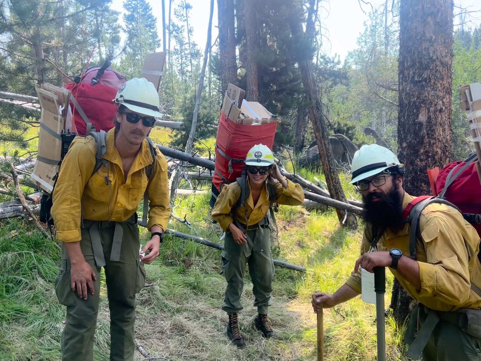 Three wildland firefighters in their firefighting apparel carry their gear on their backs.