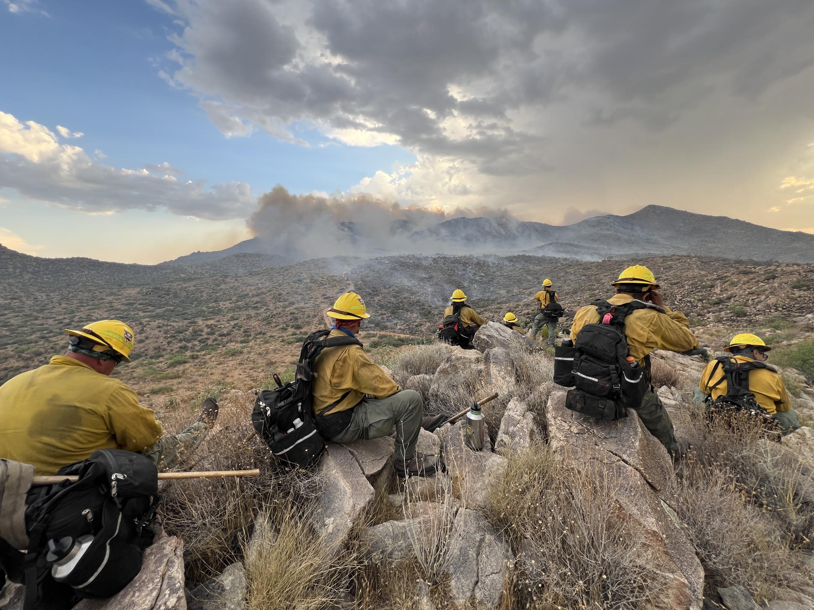 Firefighters sit on rock outcrop with smoke rising in the background