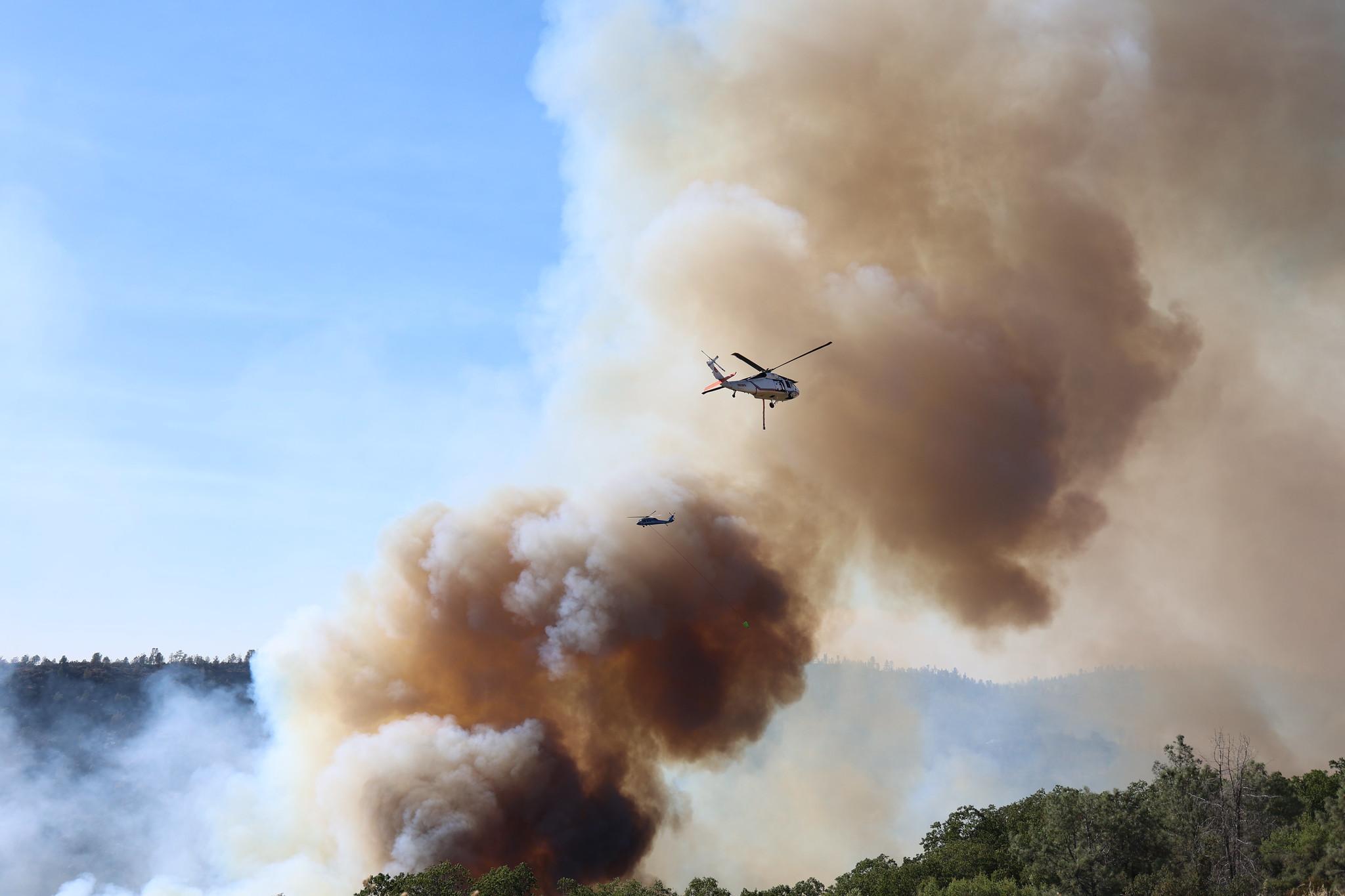 Two helicopters trailing buckets fly in opposite directions through a plume of smoke.
