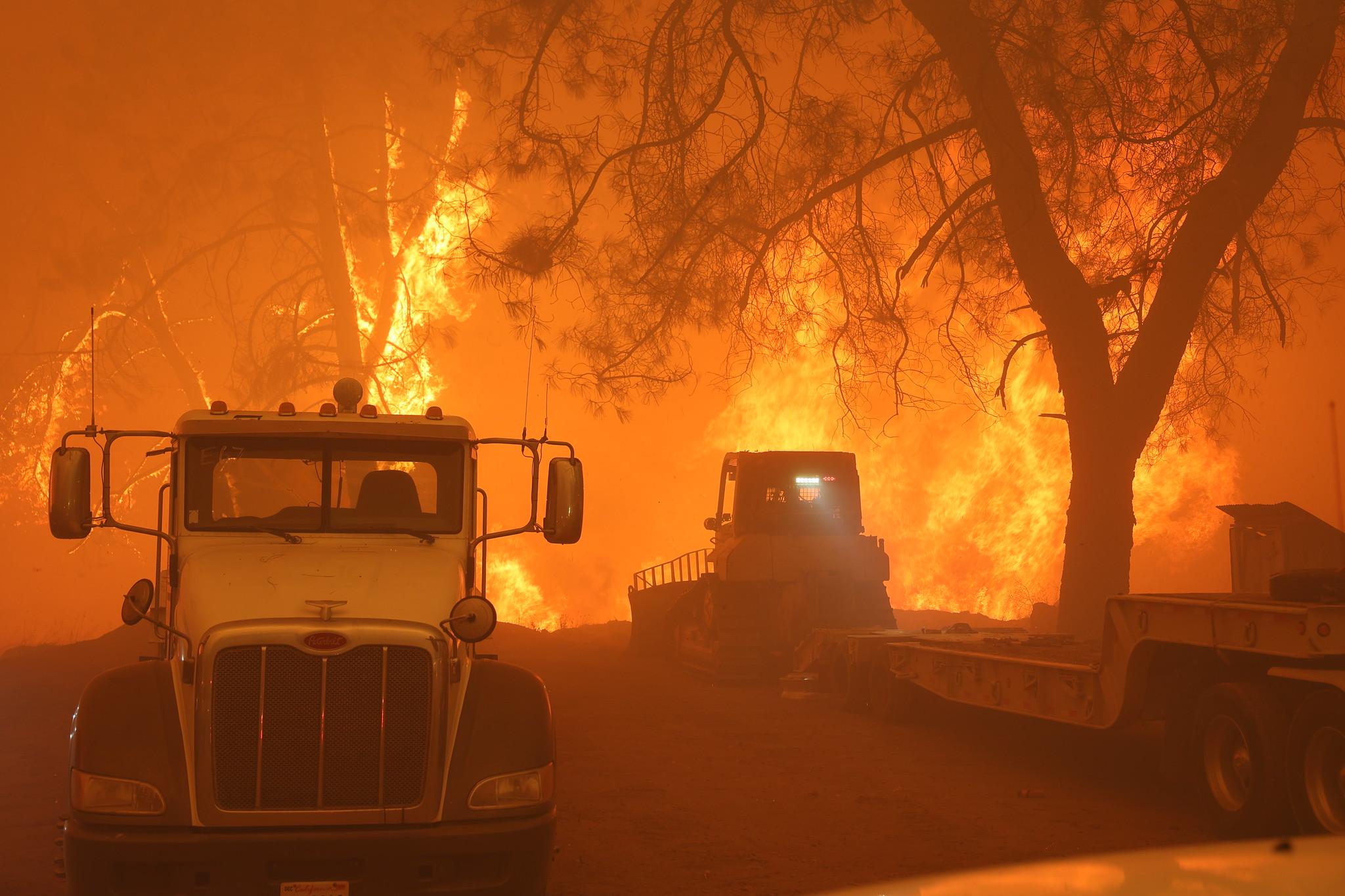 A truck and a bulldozer stand next to large orange flames.