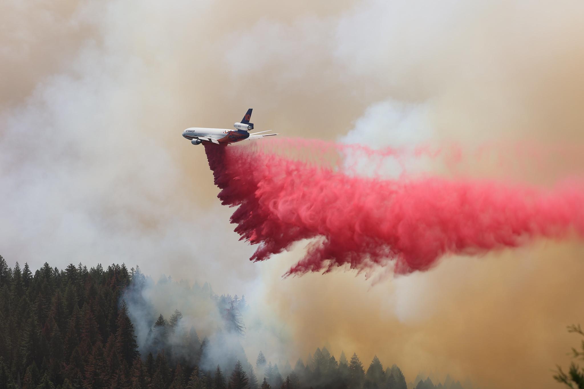 Large airplane trails red cloud of retardant through smoky skies.