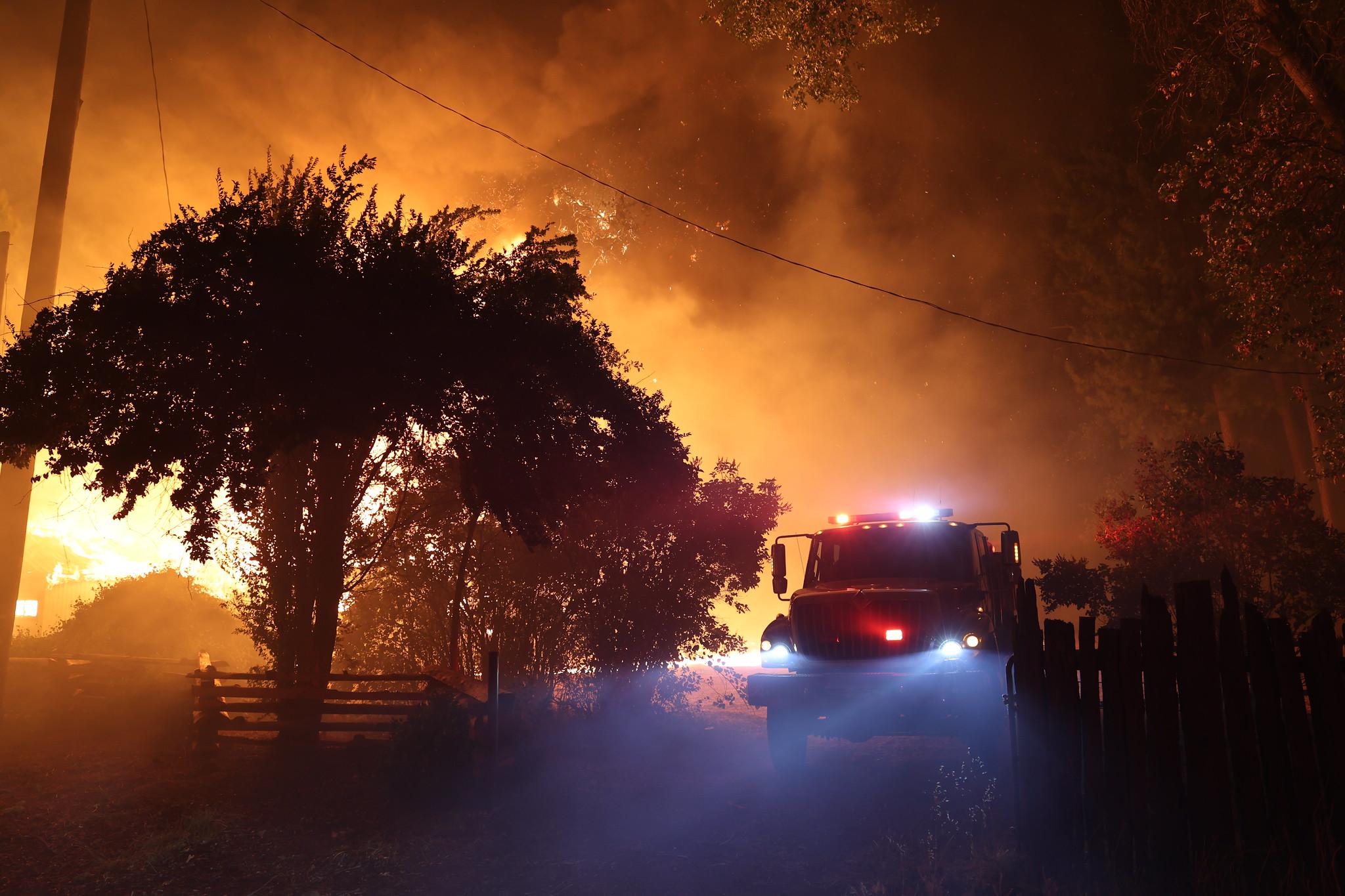 A fire engine is next to trees silhouetted by orange flames.