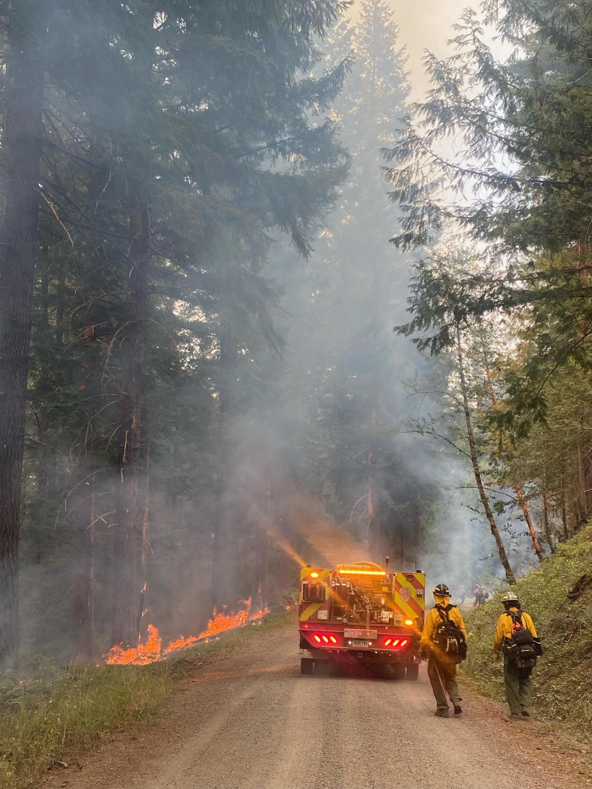 Two firefighters follow a small fire engine along a road with flames on left side of road