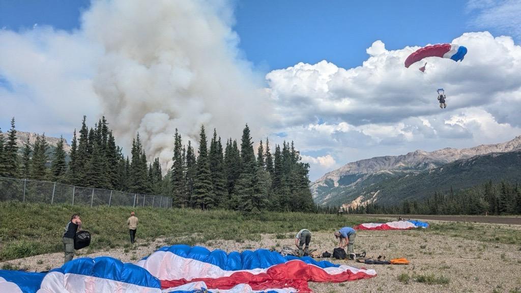A smokejumper glides in on a parachute as others on the ground stow their chutes.