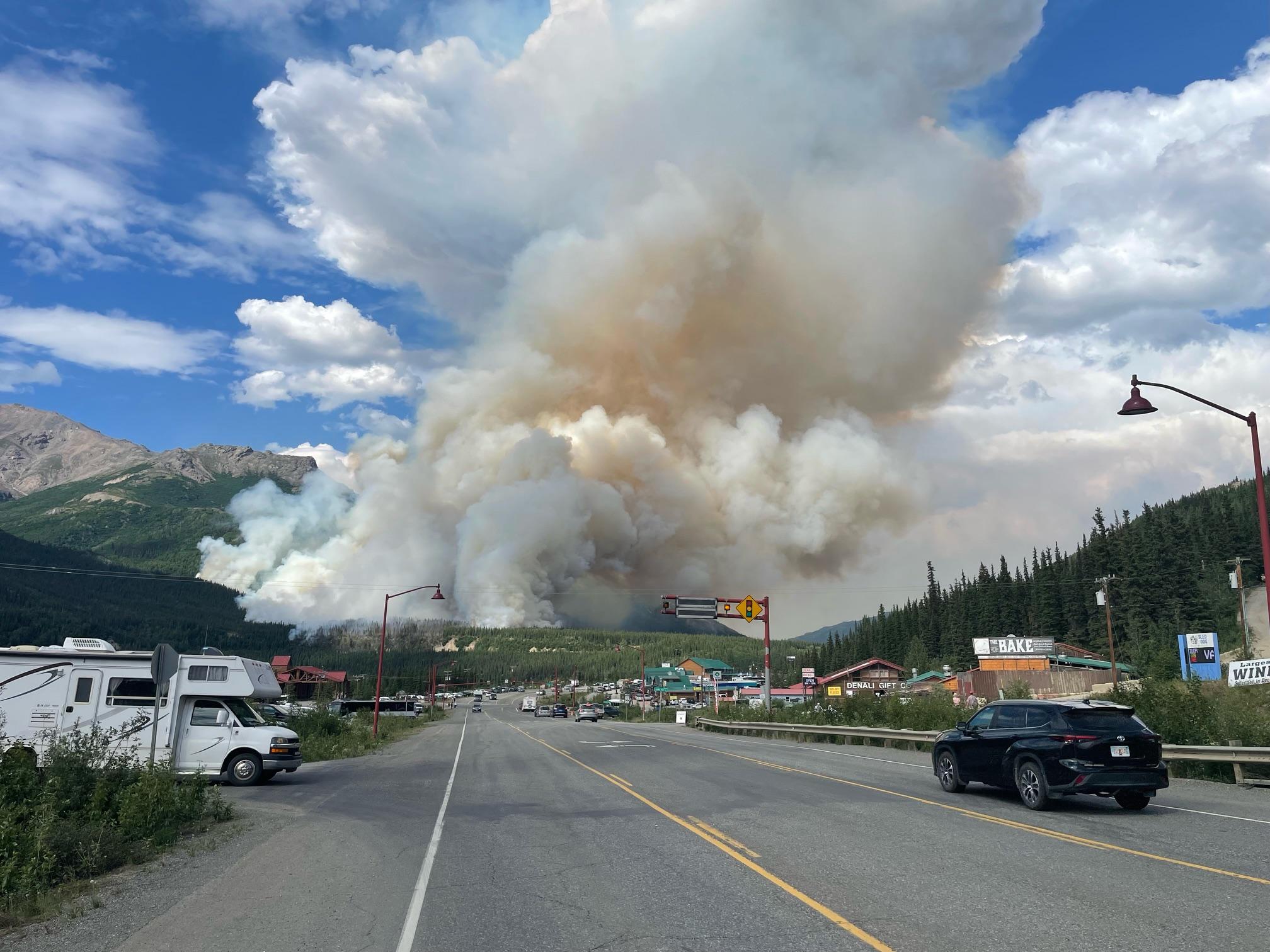 A large plume of smoke rises up the slopes of a mountain near a highway.